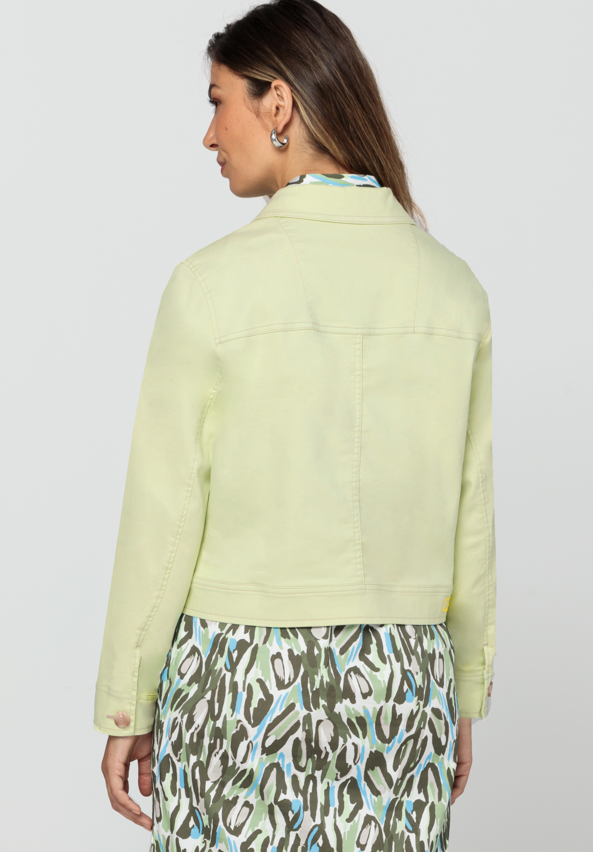 Von hinten ist eine Frau zu sehen. Sie trägt eine hellgelbe Jacke über einem gemusterten Kleid mit grünen, weißen und blauen Mustern. Sie hat langes braunes Haar und steht vor einem schlichten hellen Hintergrund.