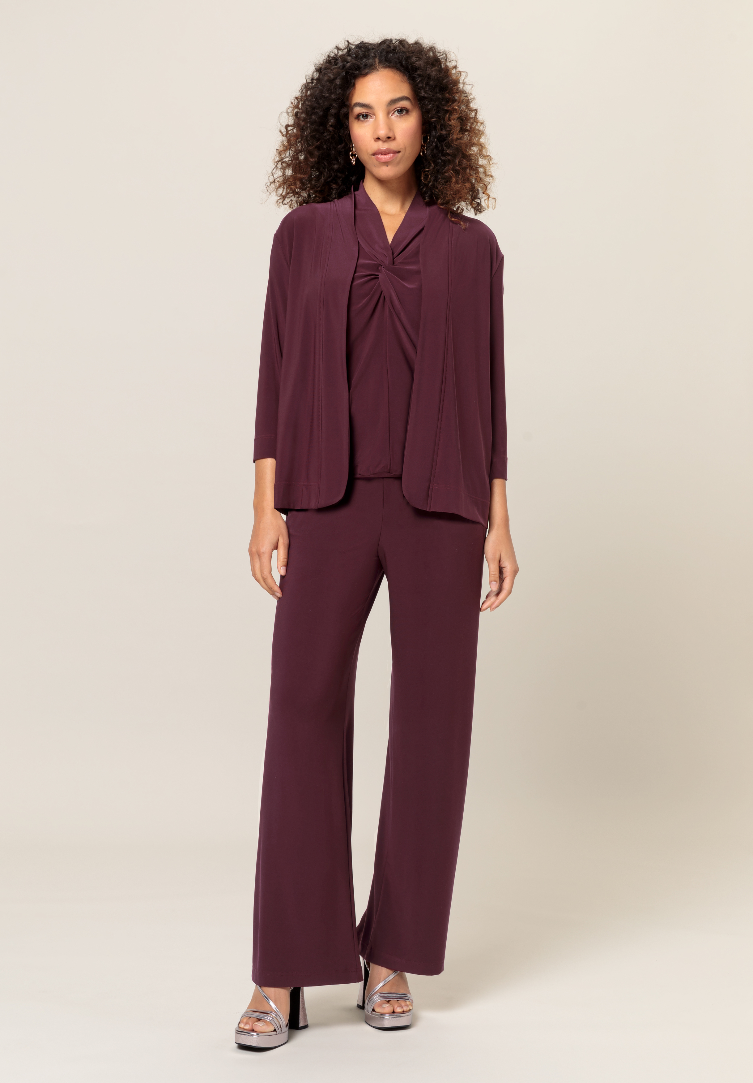Eine Frau steht vor einem schlichten hellen Hintergrund. Sie trägt ein passendes pflaumenfarbenes Outfit mit weiten Hosen, einem drapierten Oberteil und einer lockeren Strickjacke. Sie hat lockiges Haar und trägt silberne Plateausandalen.