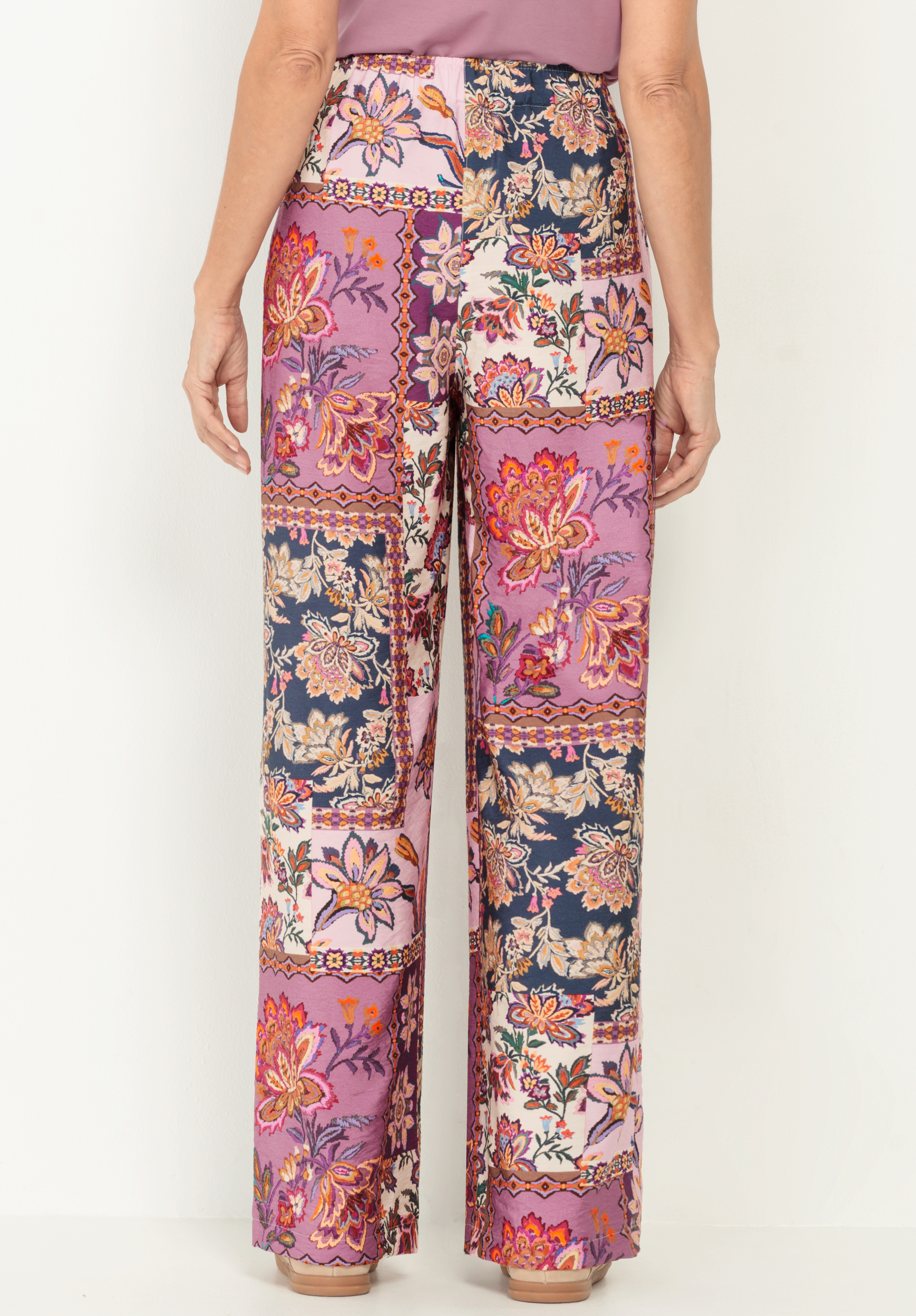 Eine Person, die eine hochtaillierte Hose mit weitem Bein und einem bunten, floralen Patchwork-Muster in Rosa-, Marine-, Orange- und Cremetönen trägt, von hinten gesehen vor einem schlichten weißen Hintergrund.