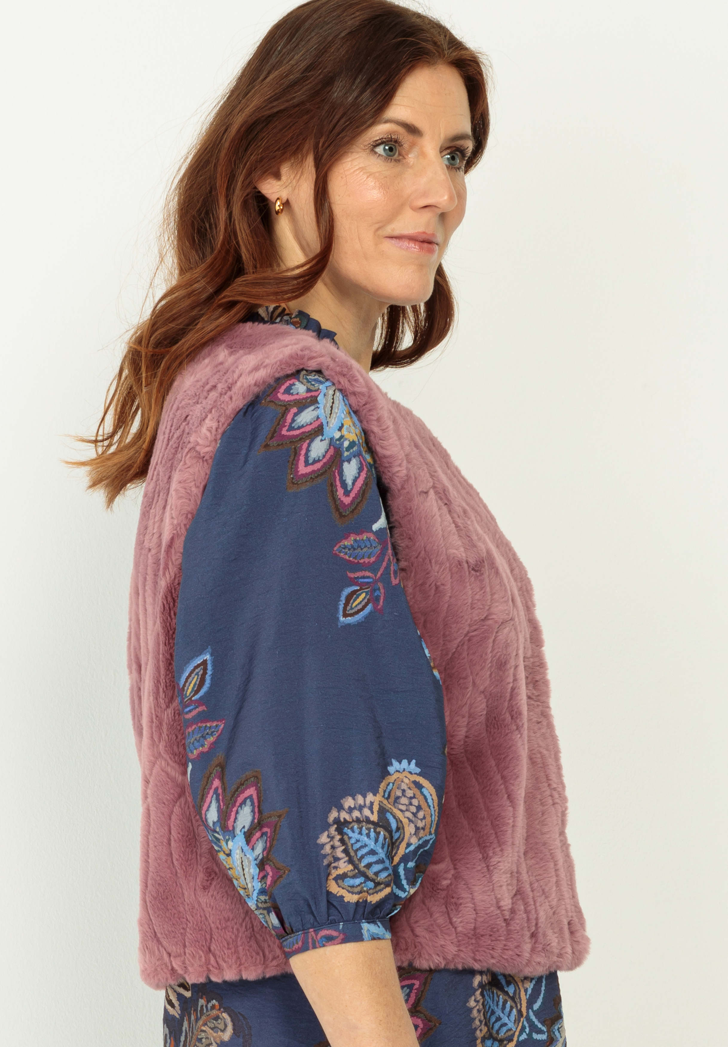 Eine Frau mit gewelltem braunem Haar trägt ein blaues Kleid mit bunten Blumenmustern und eine ärmellose, strukturierte rosa Weste. Sie steht seitwärts, nach rechts gewandt, vor einem schlichten hellen Hintergrund.