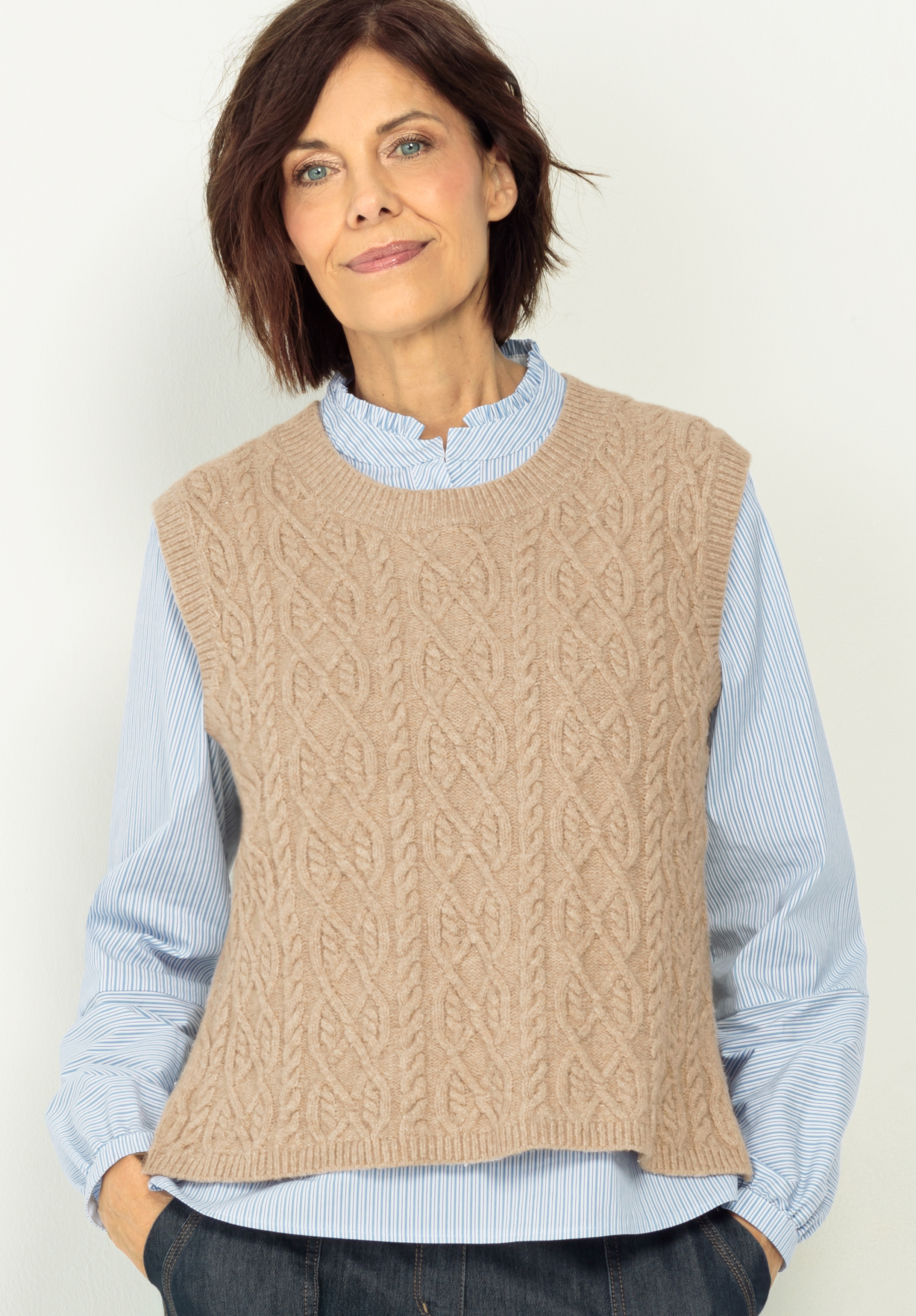 Pullunder URSY Eine Frau mit kurzen braunen Haaren trägt ein hellblaues Button-up-Hemd und einen beigen ärmellosen Pullover aus Zopfstrick. Sie steht vor einem schlichten hellen Hintergrund, blickt mit einem sanften Lächeln nach vorne und hat die Hände in den Taschen.