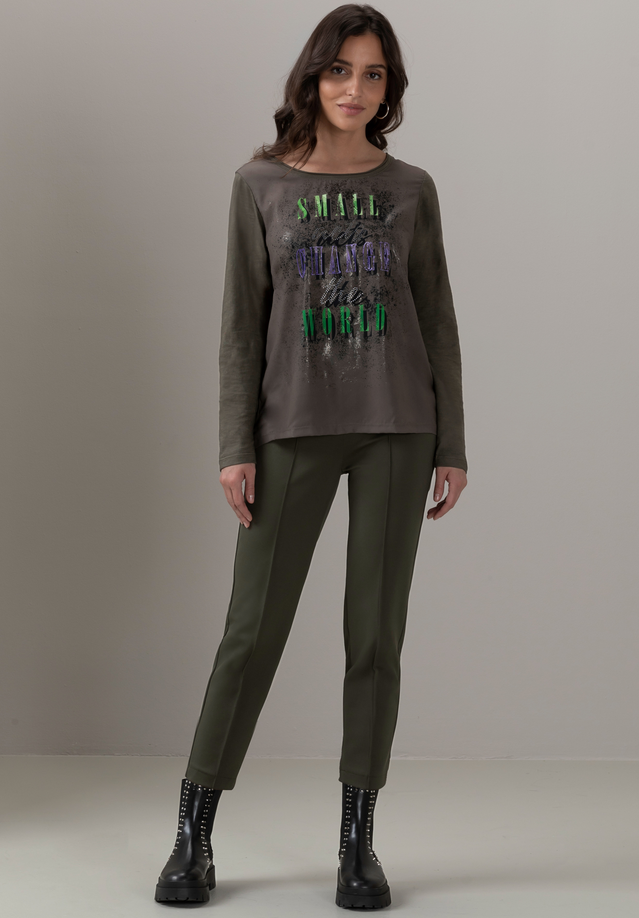 Eine Frau steht vor einem schlichten Hintergrund. Sie trägt ein olivgrünes Langarmshirt mit dem Schriftzug „SMALL CHANGE BIG WORLD“ in leuchtenden Buchstaben, dazu passende grüne Hosen und schwarze Schnürstiefel. Sie lächelt und blickt nach vorne.
