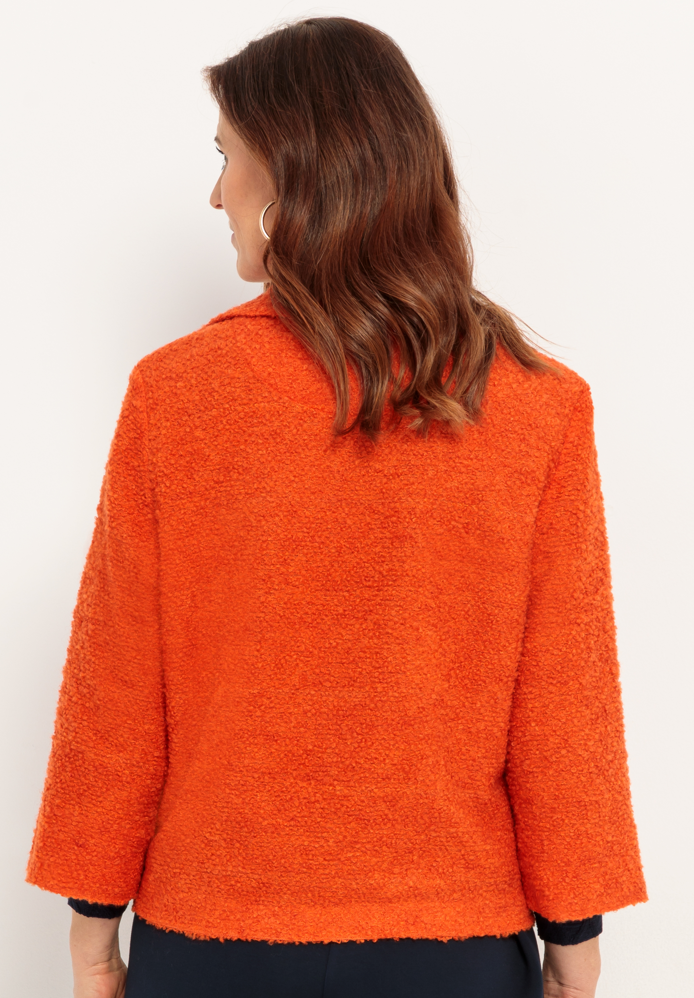 Eine Frau mit gewelltem braunem Haar ist von hinten zu sehen. Sie trägt eine strukturierte, leuchtend orangefarbene Jacke mit langen Ärmeln. Der Hintergrund ist einfarbig und hell.