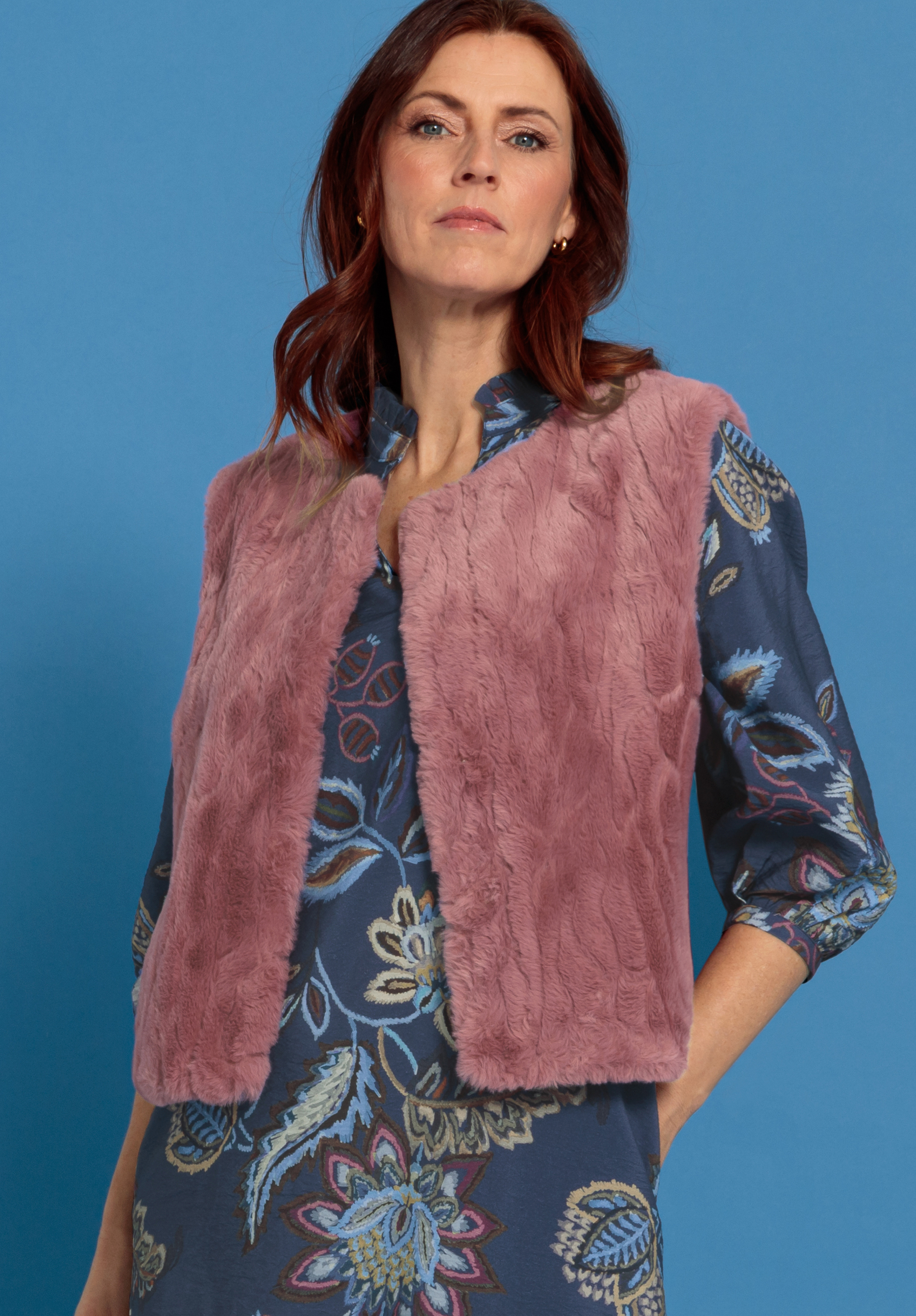 Eine Frau mit gewelltem braunem Haar, die eine lila Kunstpelzweste über einem blauen Blumenkleid trägt, steht vor einem hellblauen Hintergrund und blickt selbstbewusst in die Kamera.