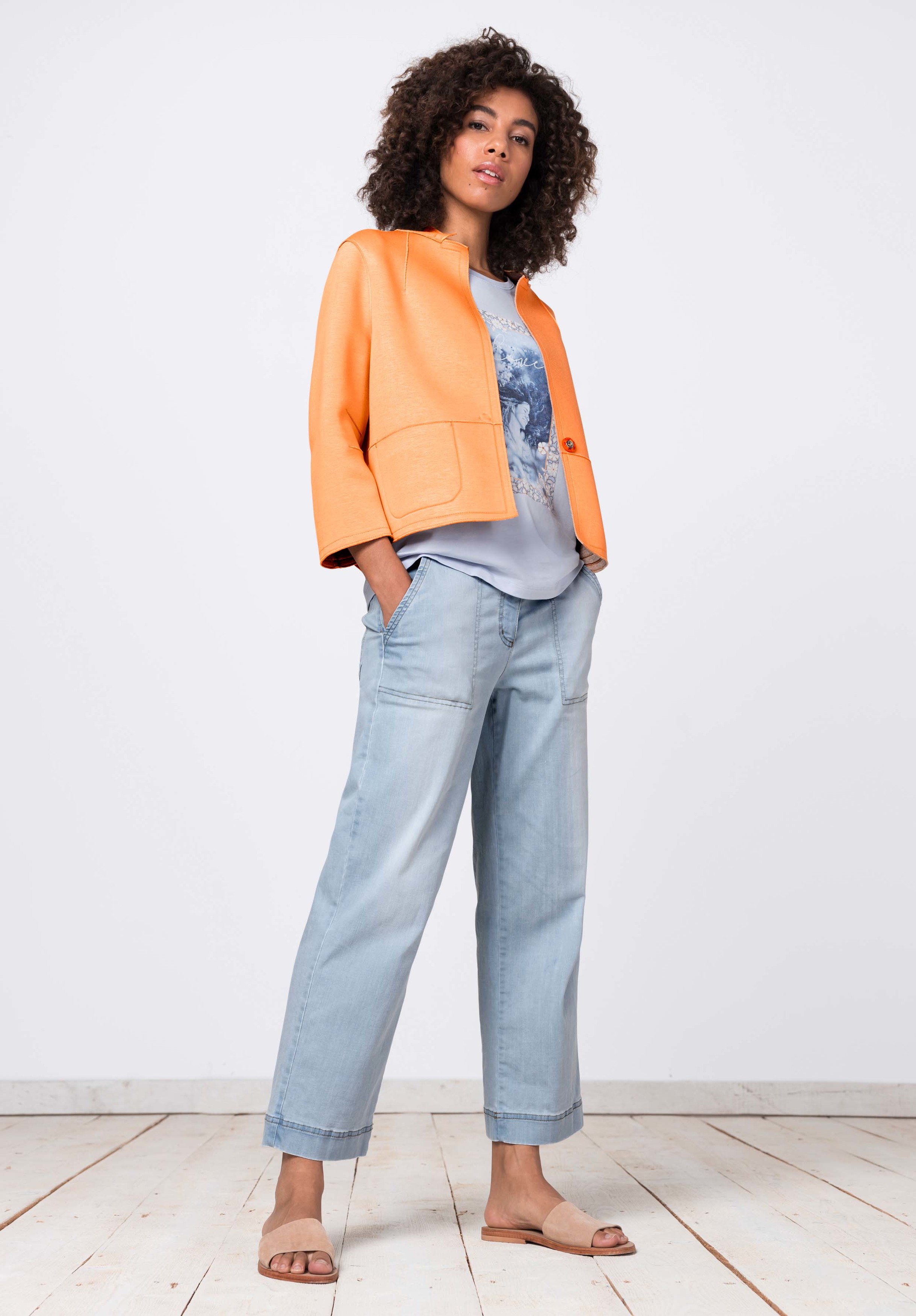 Eine Frau mit lockigem Haar steht selbstbewusst da. Sie trägt eine leuchtend orangefarbene Jacke über einem bedruckten T-Shirt, hellblaue, weite Jeans und hellbraune Sandalen und hat die Hände in den Taschen vor einem schlichten weißen Hintergrund.