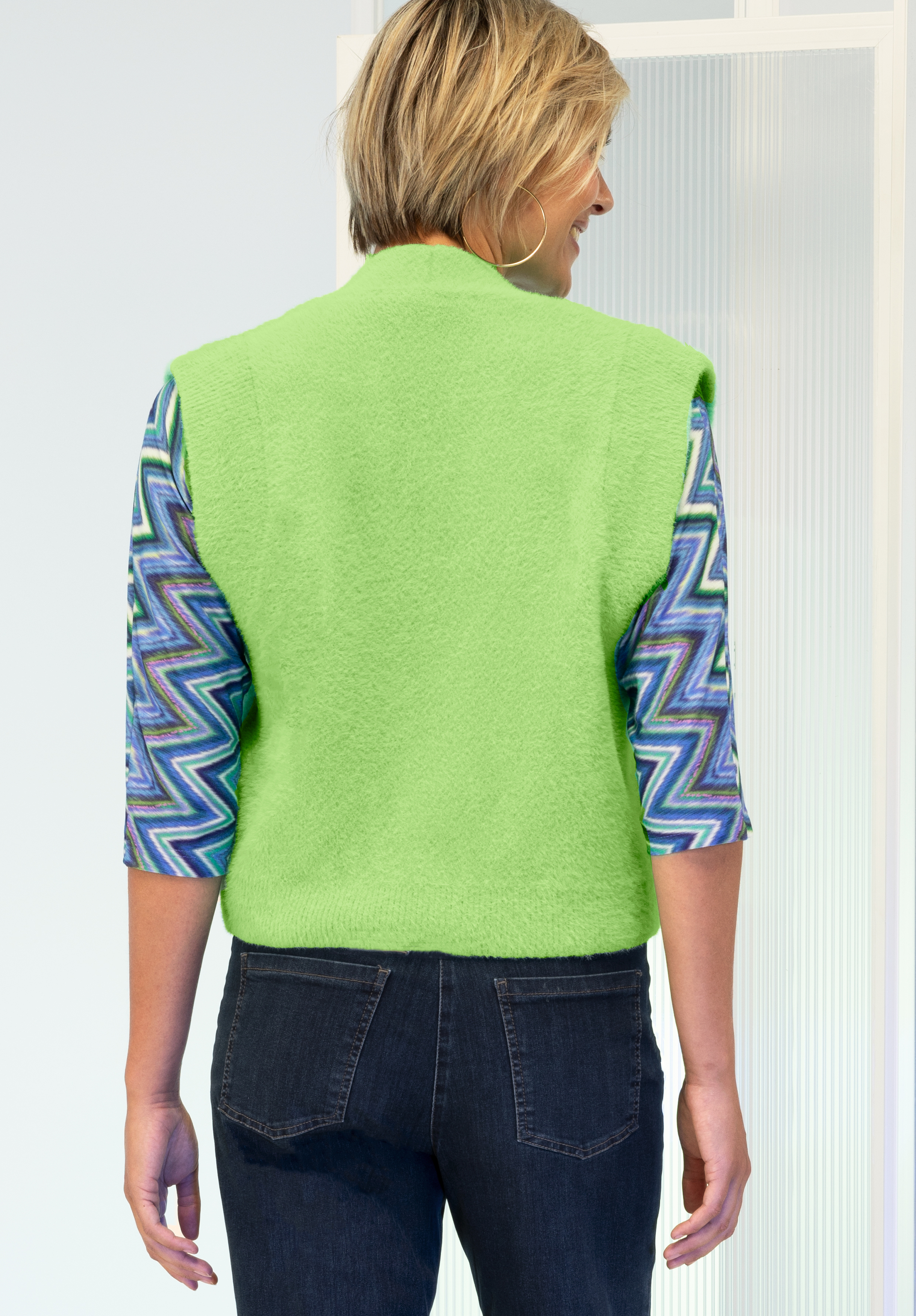 Eine Person mit kurzen blonden Haaren trägt von hinten gesehen eine hellgrüne ärmellose Weste über einem blau-violetten und weißen Langarmhemd mit Zickzackmuster und dunklen Jeans.