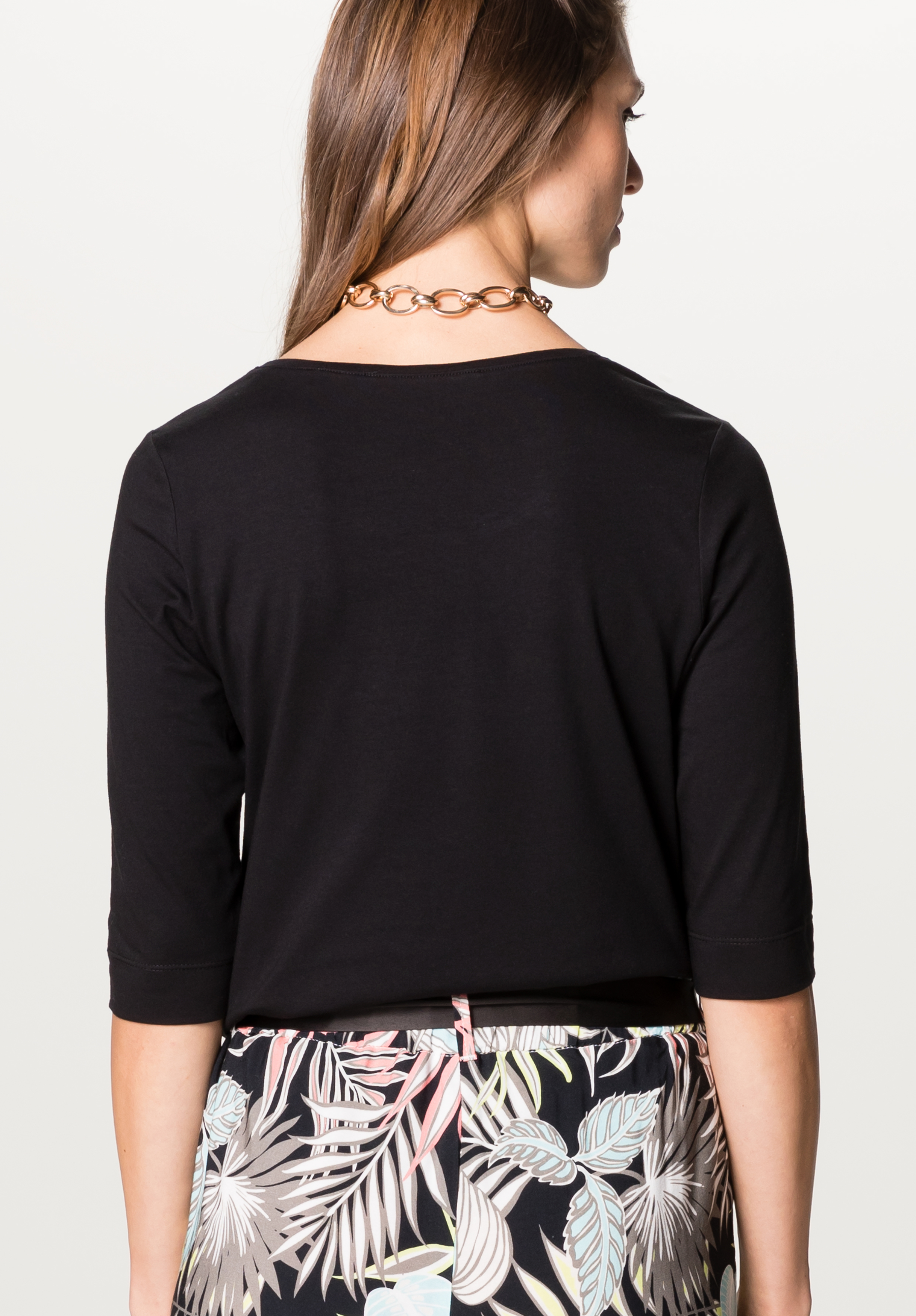 Eine Frau wird von hinten gezeigt, die ein schwarzes Oberteil mit dreiviertel Ärmeln, eine goldene Halskette und eine schwarz gemusterte Hose mit bunten Blattmustern trägt. Ihr braunes Haar ist nach unten und zu einer Seite hin offen.