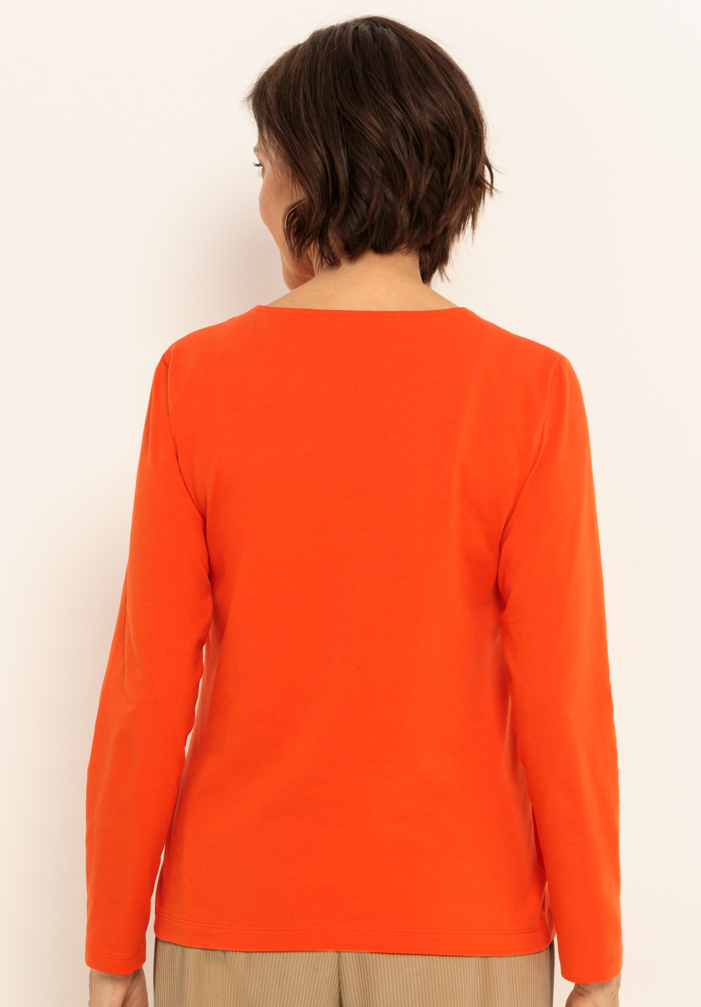 Eine Person mit kurzen braunen Haaren wird von hinten gezeigt, die ein langärmeliges, leuchtend orangefarbenes Oberteil und eine hellbraune Hose trägt und vor einem schlichten, hellen Hintergrund steht.