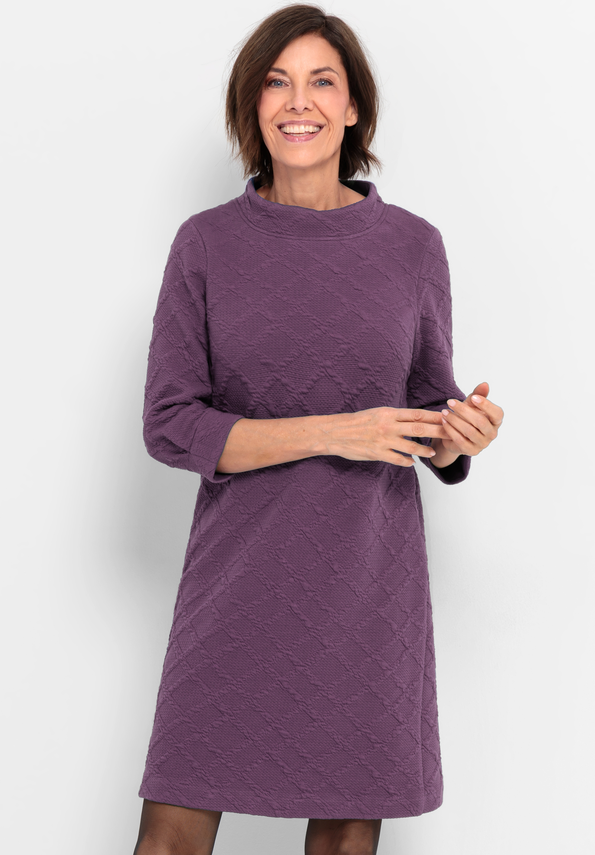 Eine Frau mit kurzen braunen Haaren lächelt, während sie ein strukturiertes, knielanges lilafarbenes Kleid mit Dreiviertelärmeln trägt, das vor einem schlichten weißen Hintergrund steht.