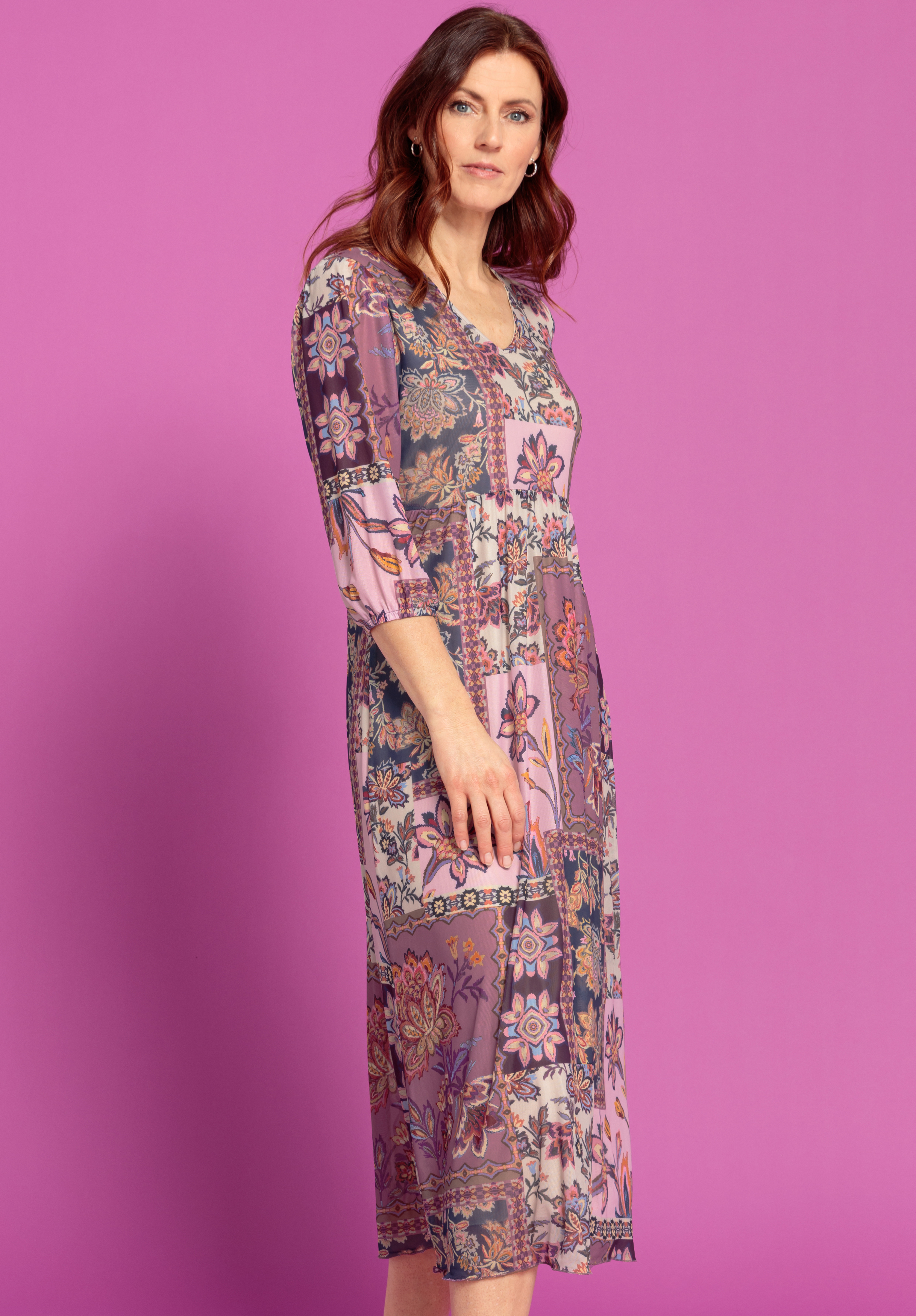 Eine Frau mit gewelltem braunem Haar, die ein buntes knielanges Kleid mit Blumenmuster und Dreiviertelärmeln trägt, steht vor einem leuchtend rosafarbenen Hintergrund, blickt leicht zur Seite und schaut in die Kamera.