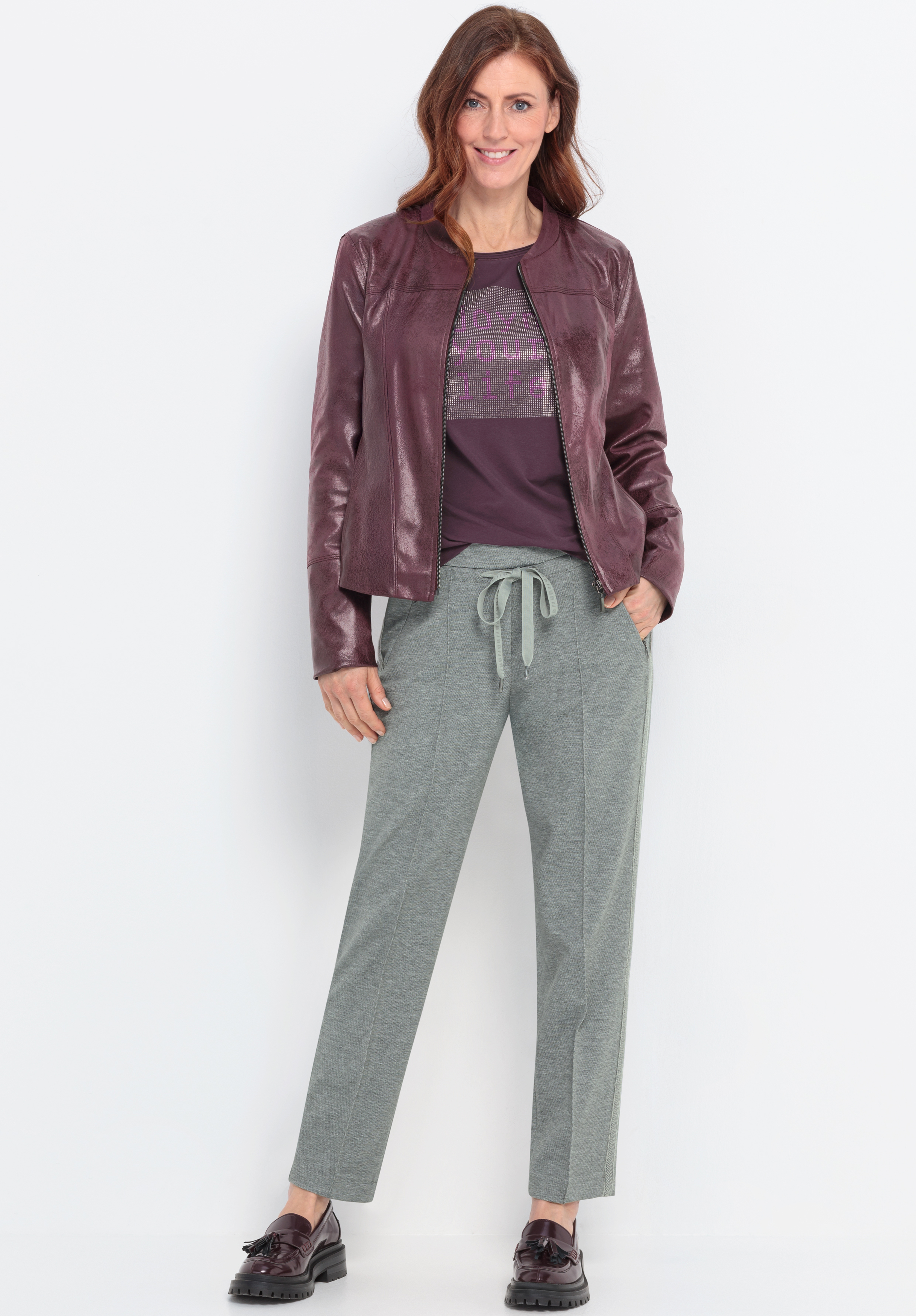 Eine Frau steht lächelnd vor einem weißen Hintergrund und trägt eine glänzende weinrote Jacke über einem lila Oberteil, eine hellgraue Hose mit Kordelzug und dunkle Halbschuhe. Ihr Haar ist gewellt und braun.