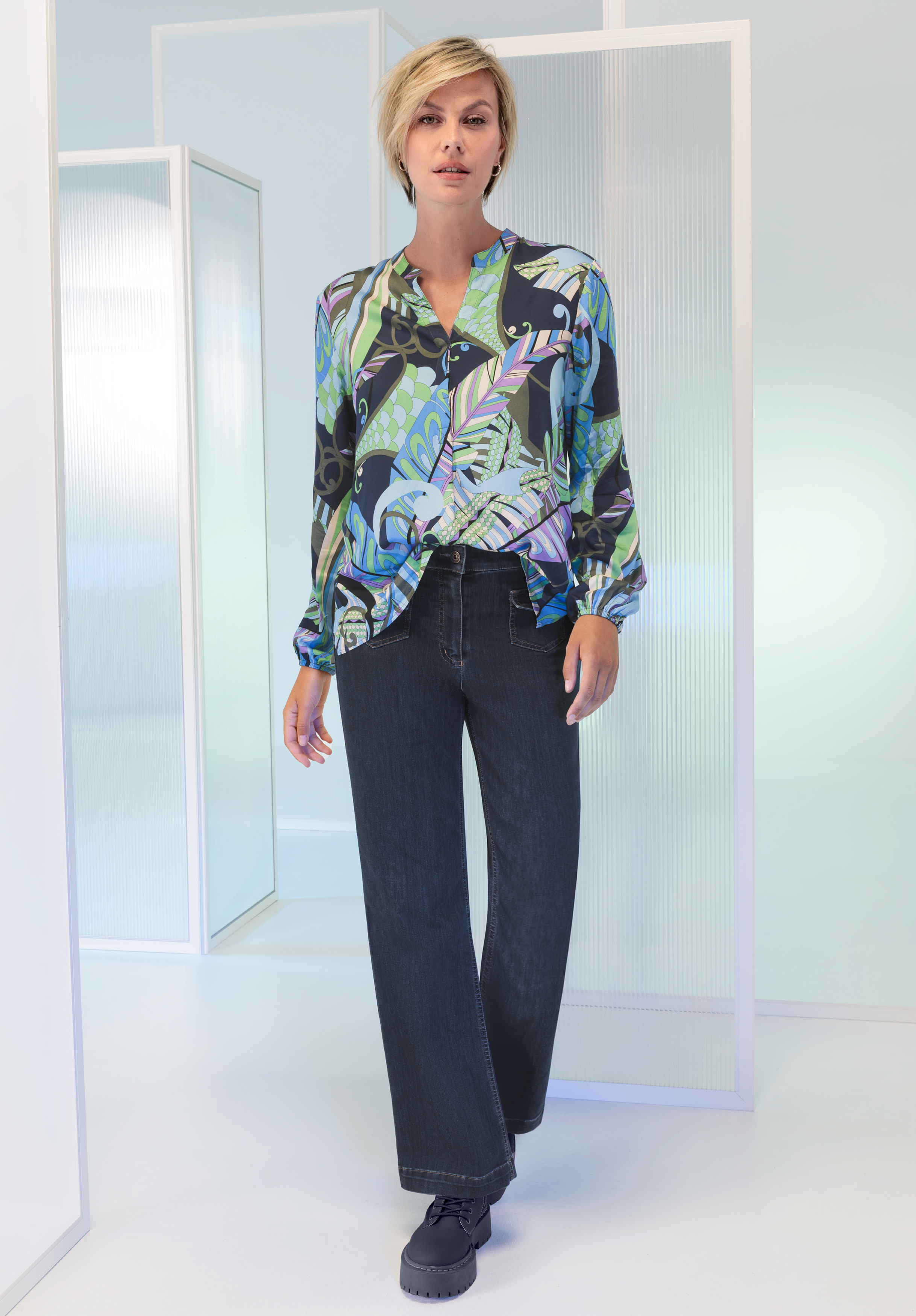Eine Frau mit kurzen blonden Haaren trägt eine bunte, gemusterte Bluse in Blau-, Grün- und Schwarztönen, dazu dunkle Jeans mit weitem Bein und dunkle Schuhe. Sie steht in einer minimalistischen, modernen Umgebung mit weißen Paneelen.