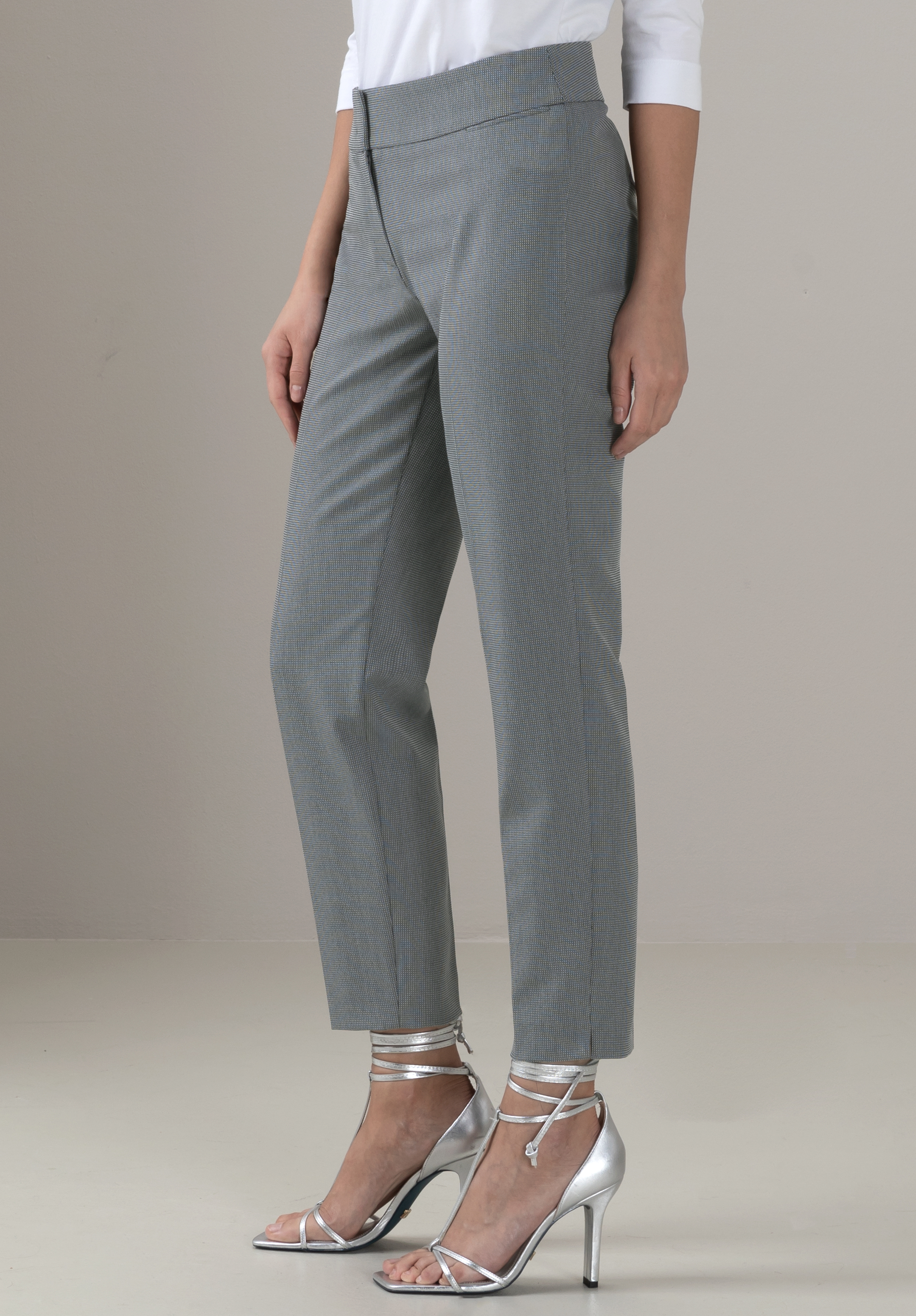 Eine Person, die eine graue maßgeschneiderte Hose, ein weißes langärmeliges Oberteil und metallisch-silberne hochhackige Sandalen mit Knöchelriemen trägt, steht vor einem schlichten hellen Hintergrund.