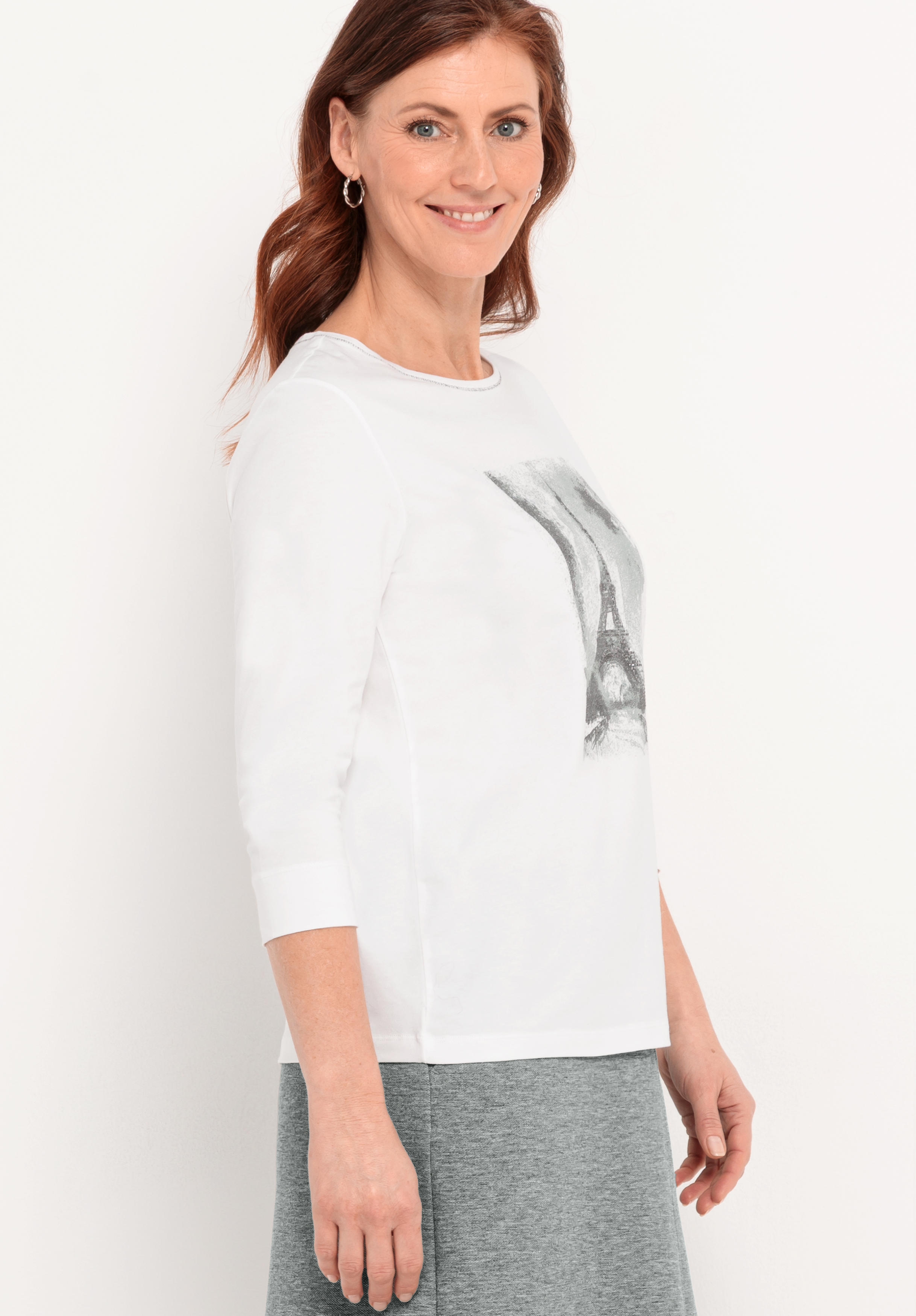 Eine Frau mit langen roten Haaren, die ein weißes dreiviertelärmeliges Oberteil mit einer Grafik des Eiffelturms und einen grauen Rock trägt, steht lächelnd und leicht zur Seite gewandt vor einem unifarbenen Hintergrund.