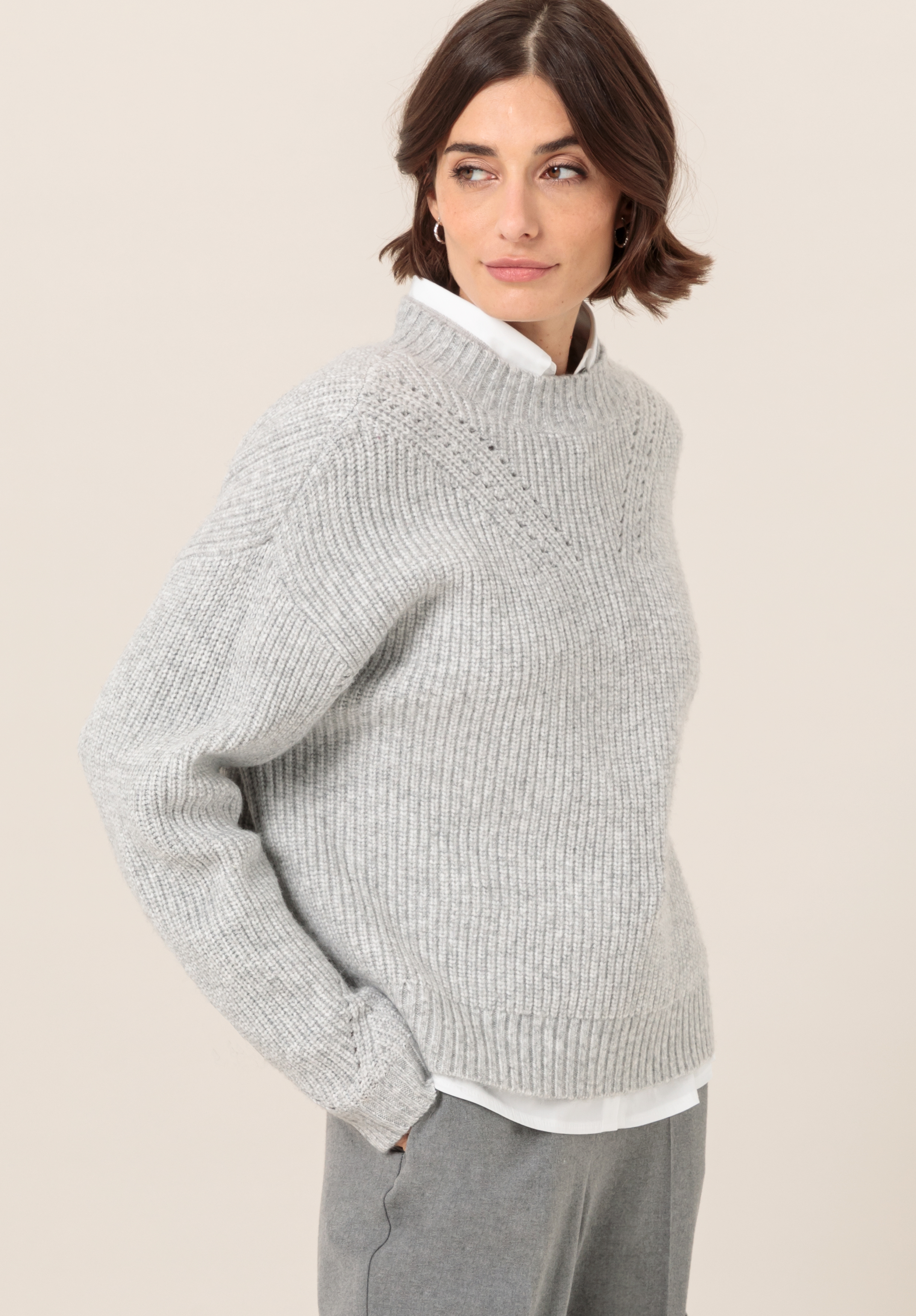 Eine Frau mit kurzen braunen Haaren trägt einen hellgrauen Strickpullover über einem weißen Hemd mit Kragen und einer grauen Hose. Sie steht mit den Händen in den Taschen da und blickt zur Seite. Der Hintergrund ist schlicht und hell.
