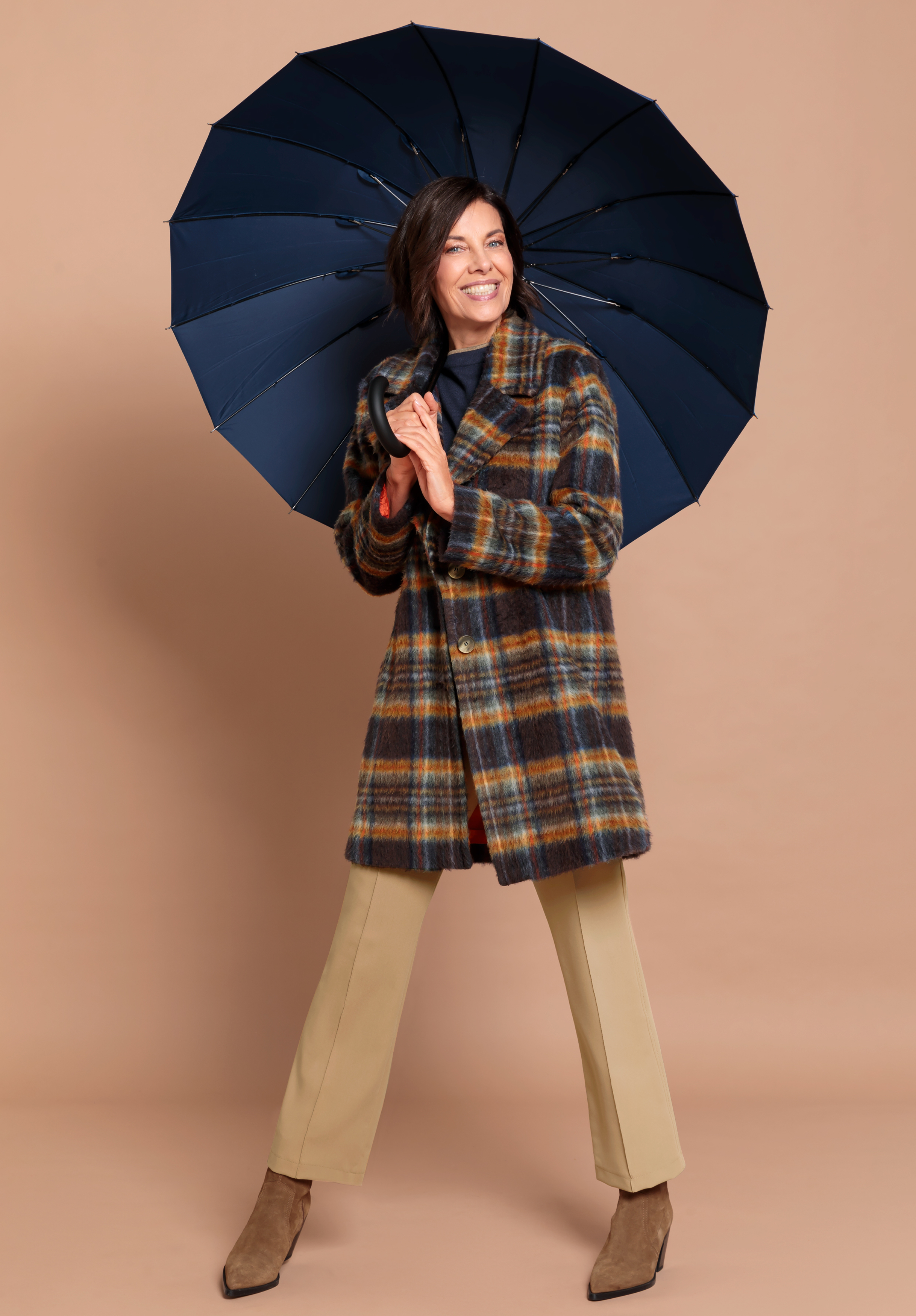 Eine Frau lächelt, während sie einen dunkelblauen Regenschirm hält. Sie trägt einen braun und blau karierten Mantel, eine beigefarbene Hose und braune Stiefel und steht vor einem einfarbig beigen Hintergrund.