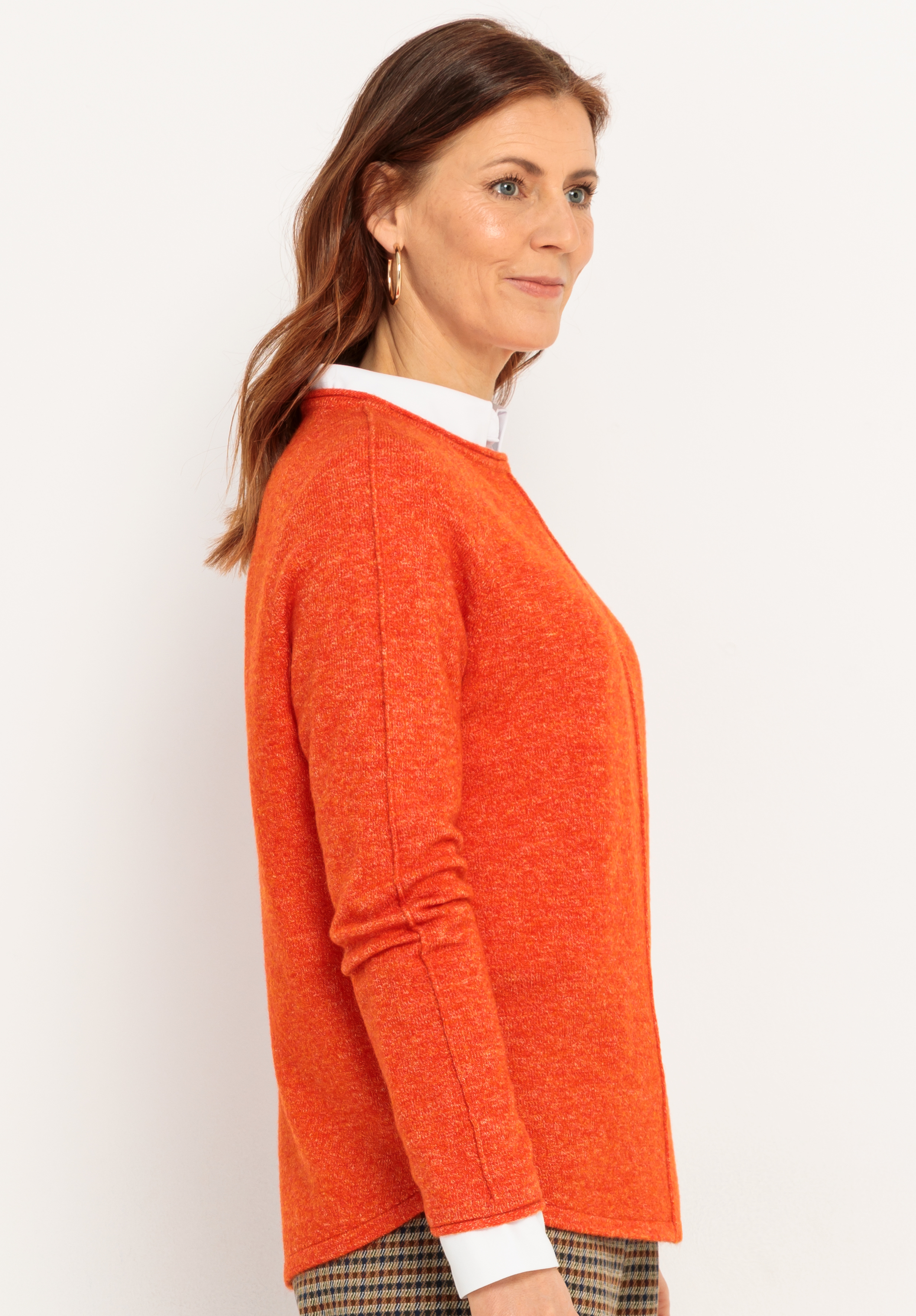 Eine Frau mit braunem Haar steht im Profil, trägt einen leuchtend orangefarbenen Pullover über einem weißen Hemd mit Kragen und eine karierte Hose vor einem schlichten weißen Hintergrund. Sie blickt leicht nach vorne und lächelt sanft.