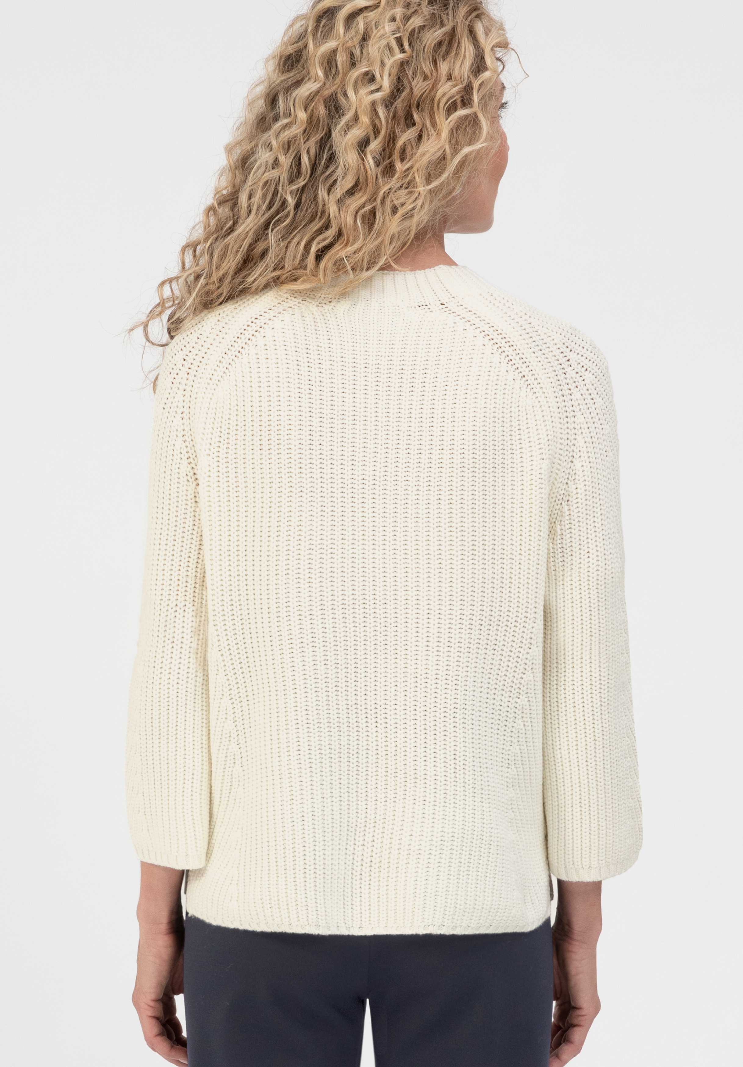 Vor einem schlichten weißen Hintergrund ist eine Person mit langen, lockigen blonden Haaren von hinten zu sehen, die einen cremefarbenen Strickpullover mit Dreiviertelärmeln und eine dunkle Hose trägt.