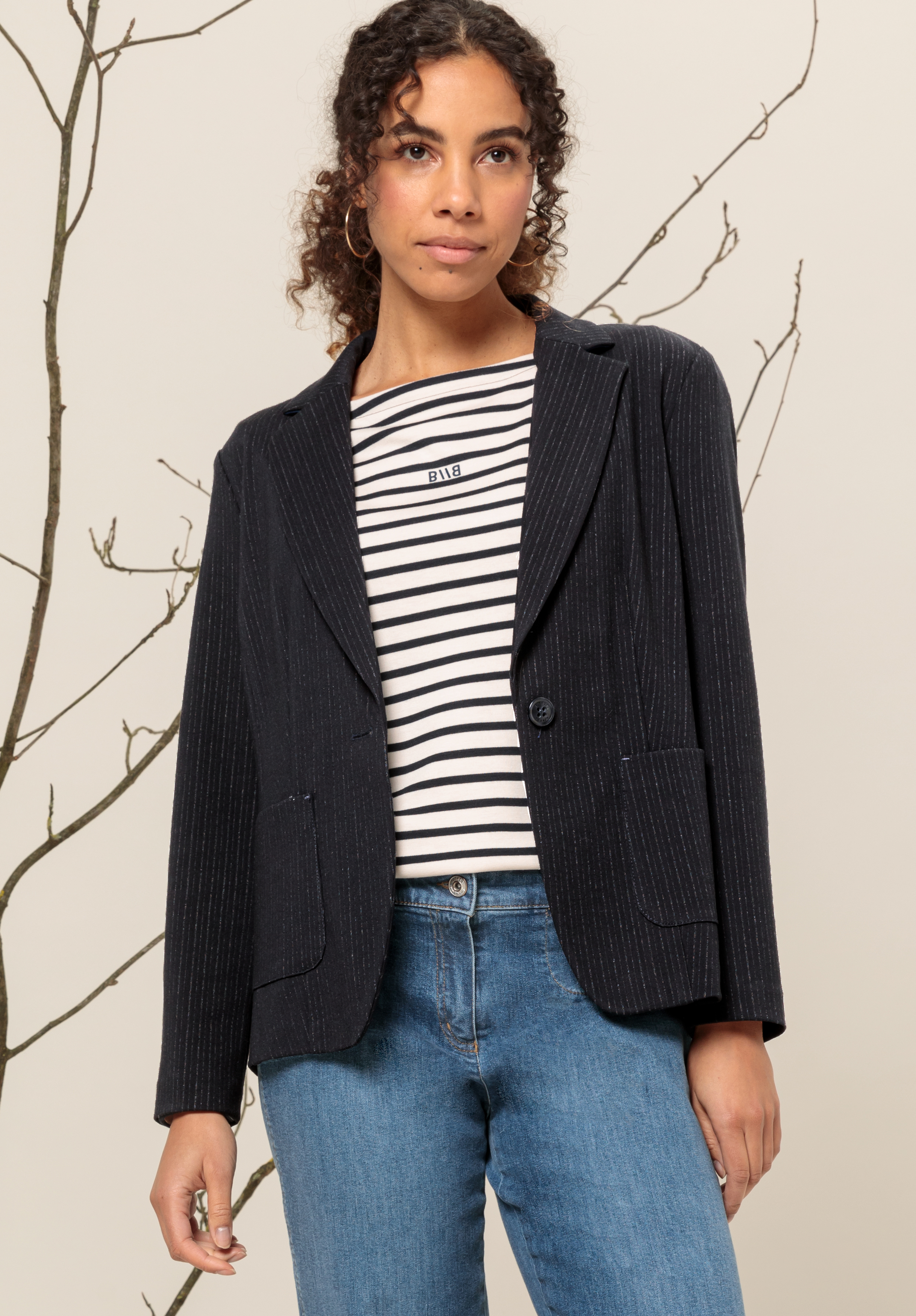 Eine Frau mit lockigem Haar, die einen schwarzen Nadelstreifenblazer, ein schwarz-weiß gestreiftes Hemd und Bluejeans trägt, steht vor einem beigen Hintergrund mit kahlen Ästen.