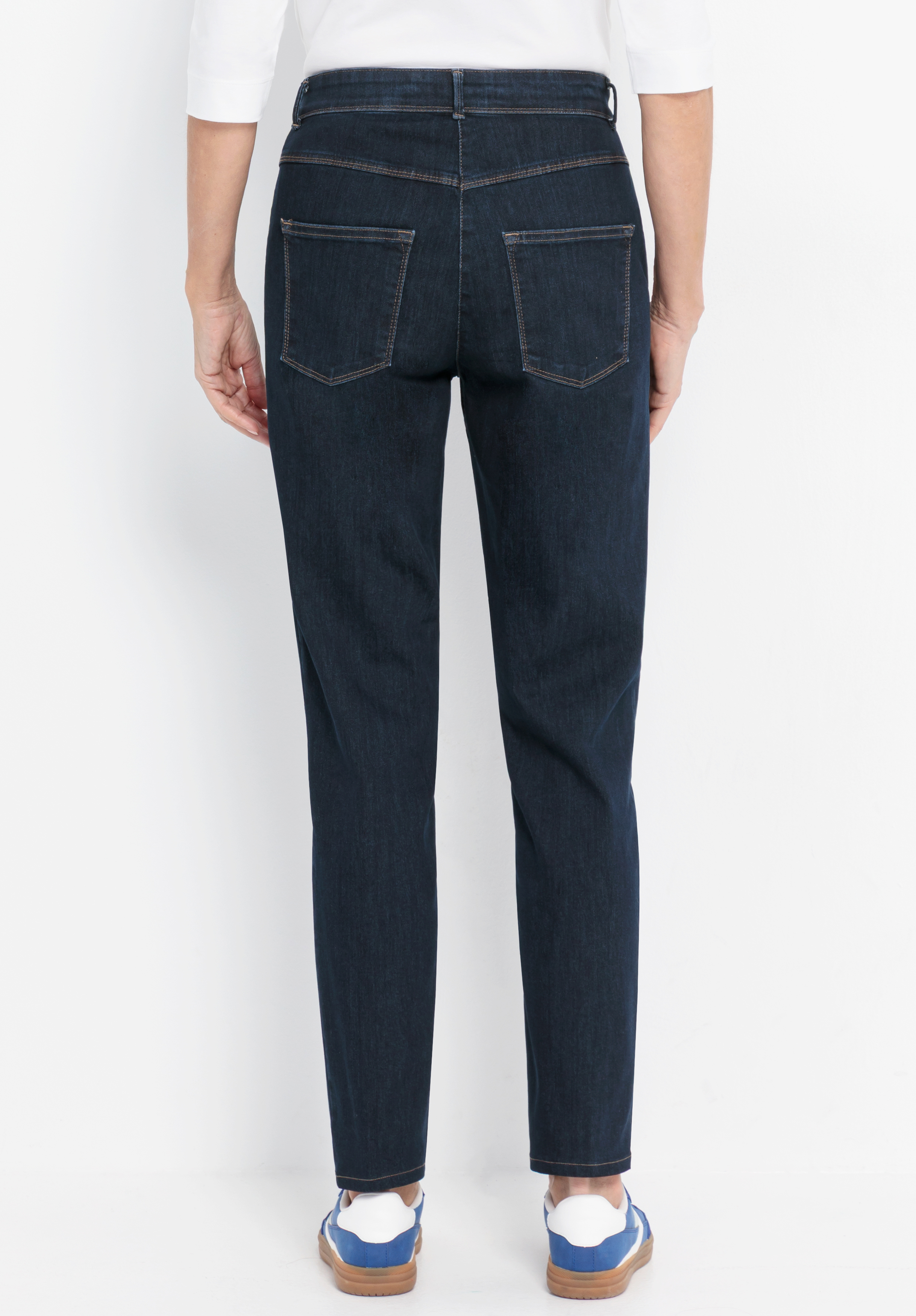 Eine Person steht mit dem Rücken zur Kamera und trägt dunkelblaue Jeans mit sichtbaren Gesäßtaschen, ein weißes Oberteil mit Ärmeln bis zu den Ellbogen sowie blau-weiße Turnschuhe. Der Hintergrund ist einfarbig weiß.