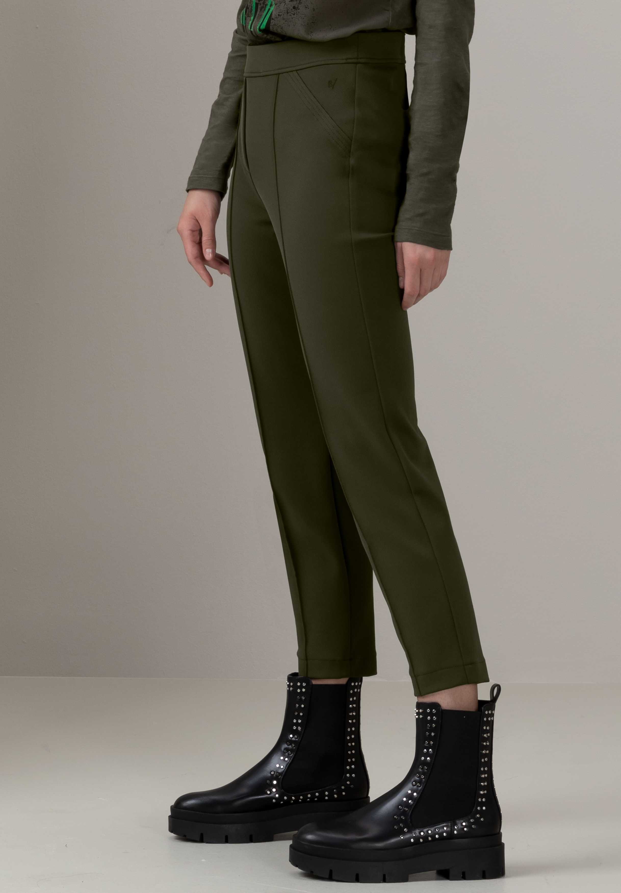 Eine Person in hoch taillierten, olivgrünen Hosen und einem langärmligen Oberteil steht vor einem neutralen Hintergrund. Sie trägt schwarze Stiefeletten mit Nietendetails. Der Kopf ist nicht sichtbar.