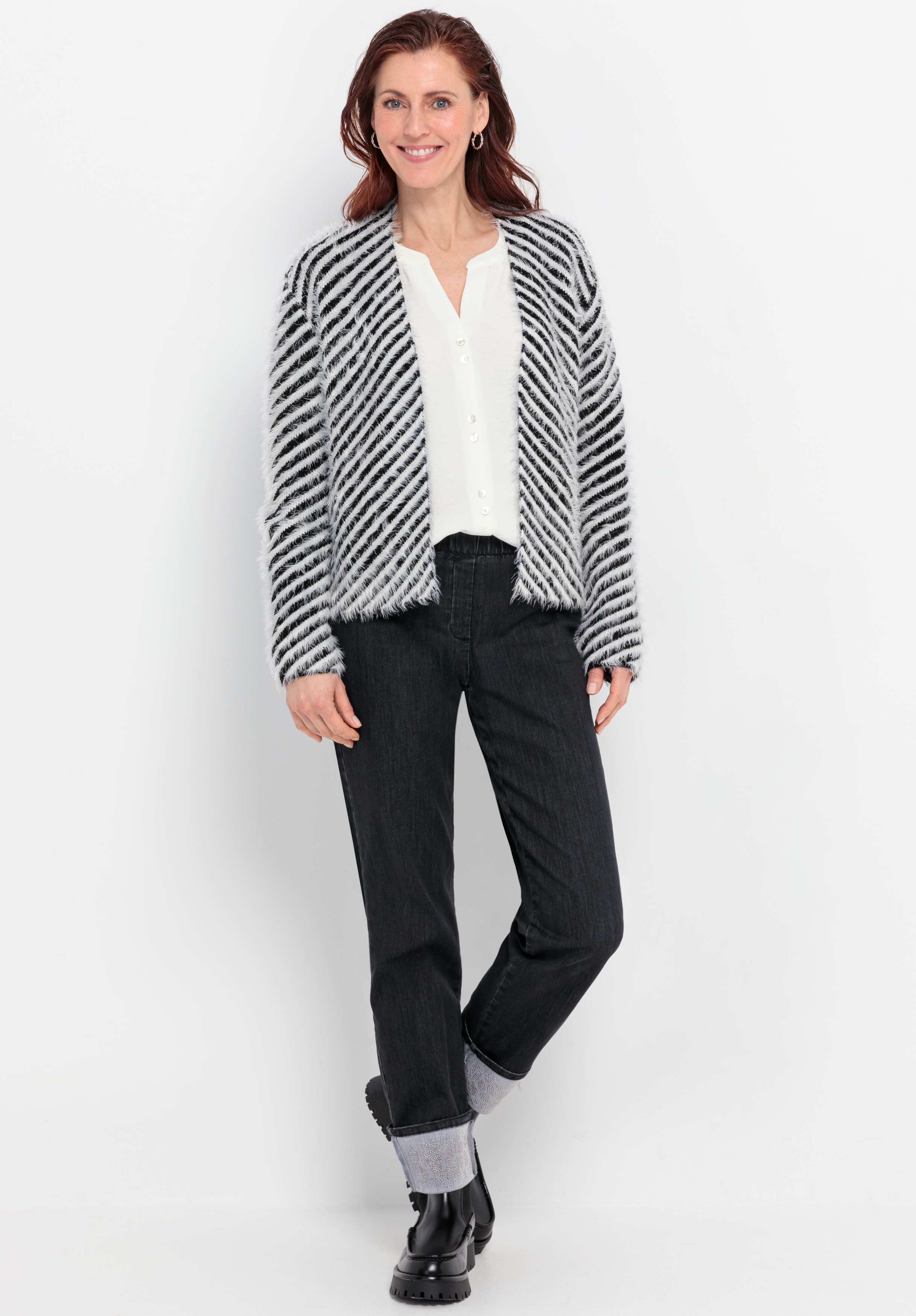 Vor einem weißen Hintergrund steht eine lächelnde Frau, die eine schwarz-weiß gestreifte Strickjacke, eine weiße Bluse, eine schwarze Jeans mit Umschlag und schwarze Stiefel trägt.
