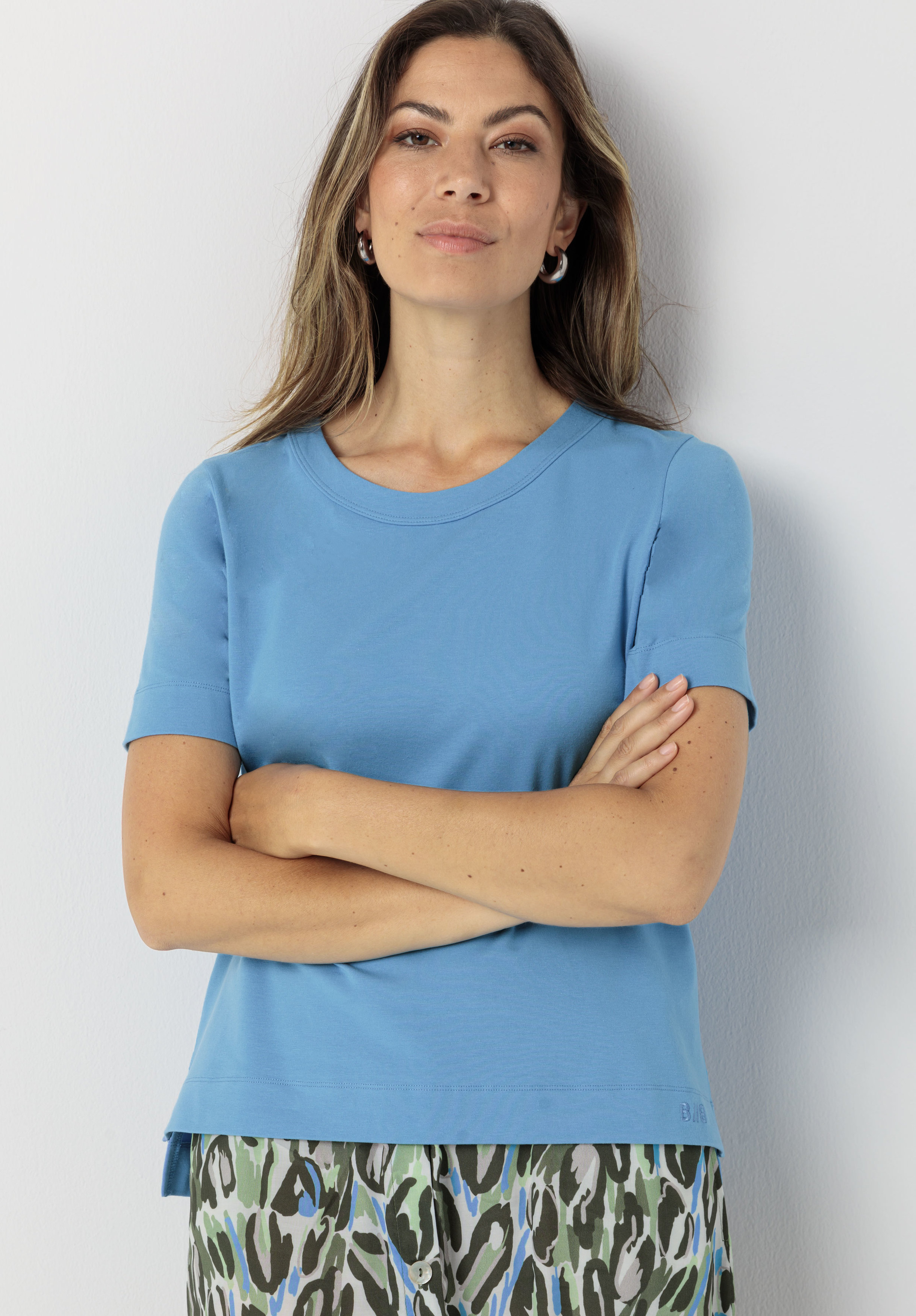 Frau mit langen Haaren, hellblauem Kurzarmshirt und gemustertem Rock steht mit verschränkten Armen vor einer hellen Wand und blickt selbstbewusst in die Kamera.