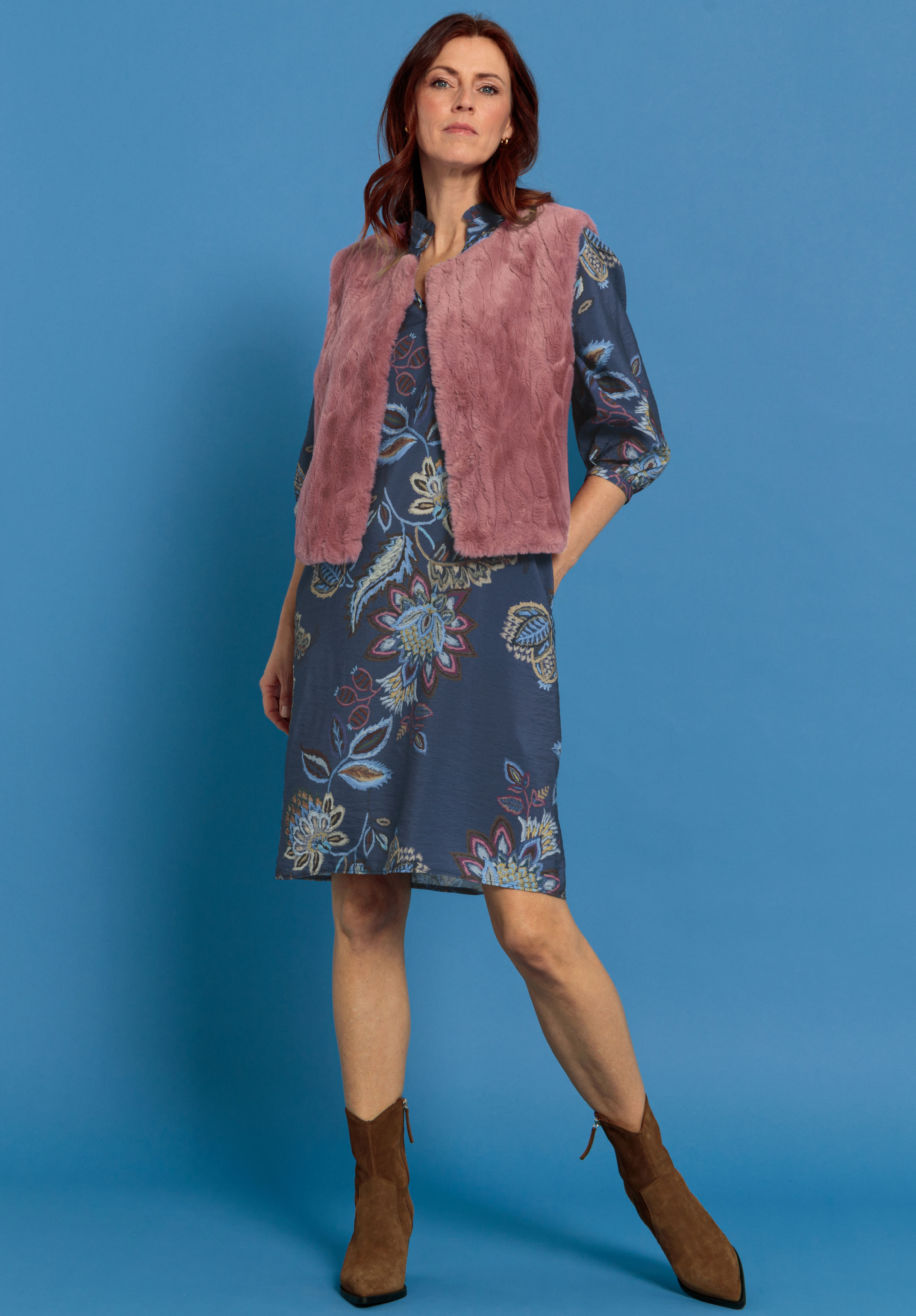 Eine Frau steht vor einem blauen Hintergrund und trägt ein blaues geblümtes Kleid, eine rosa Kunstpelzweste und braune Wildlederstiefel. Sie hat eine Hand in der Tasche und einen selbstbewussten Gesichtsausdruck.