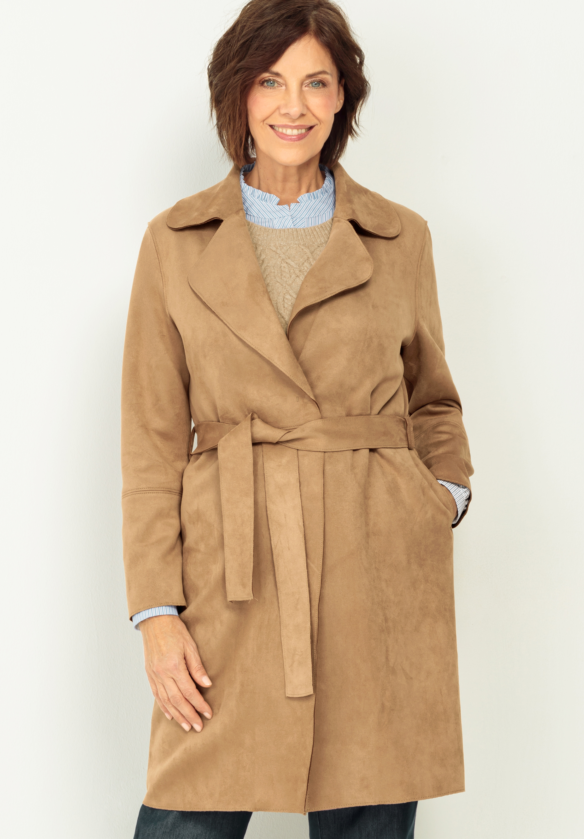 Lederimitatmantel VIANNE Eine Frau mit kurzen braunen Haaren lächelt, während sie einen hellbraunen Trenchcoat mit Gürtel über einem blauen Hemd mit Kragen und einem beigen Pullover vor einem schlichten hellen Hintergrund trägt.
