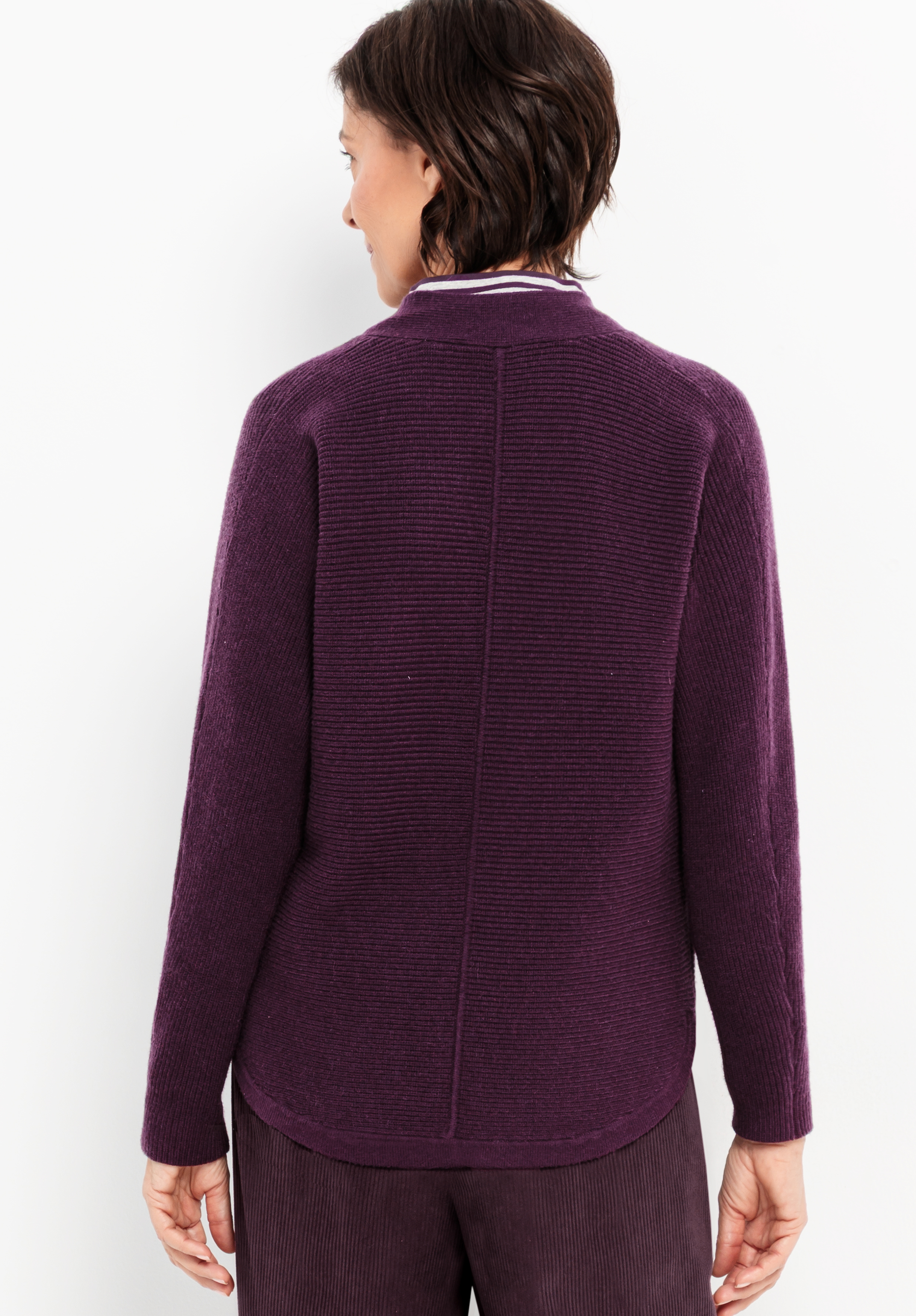 Eine Frau mit kurzen dunklen Haaren steht mit dem Rücken zur Kamera und trägt einen strukturierten, langärmeligen, dunkelvioletten Pullover und eine passende Hose vor einem schlichten weißen Hintergrund.
