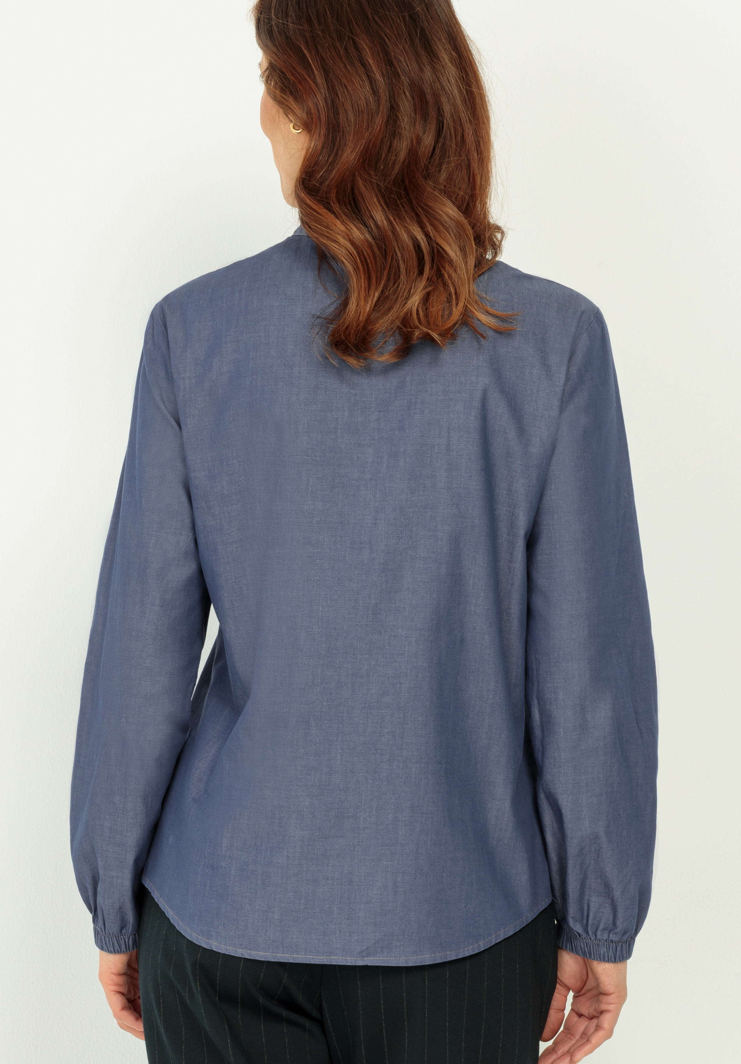 Eine Frau mit gewelltem braunem Haar ist von hinten zu sehen. Sie trägt ein langärmeliges, blaues Button-up-Hemd und eine dunkle Hose. Sie steht vor einem schlichten, hellen Hintergrund.