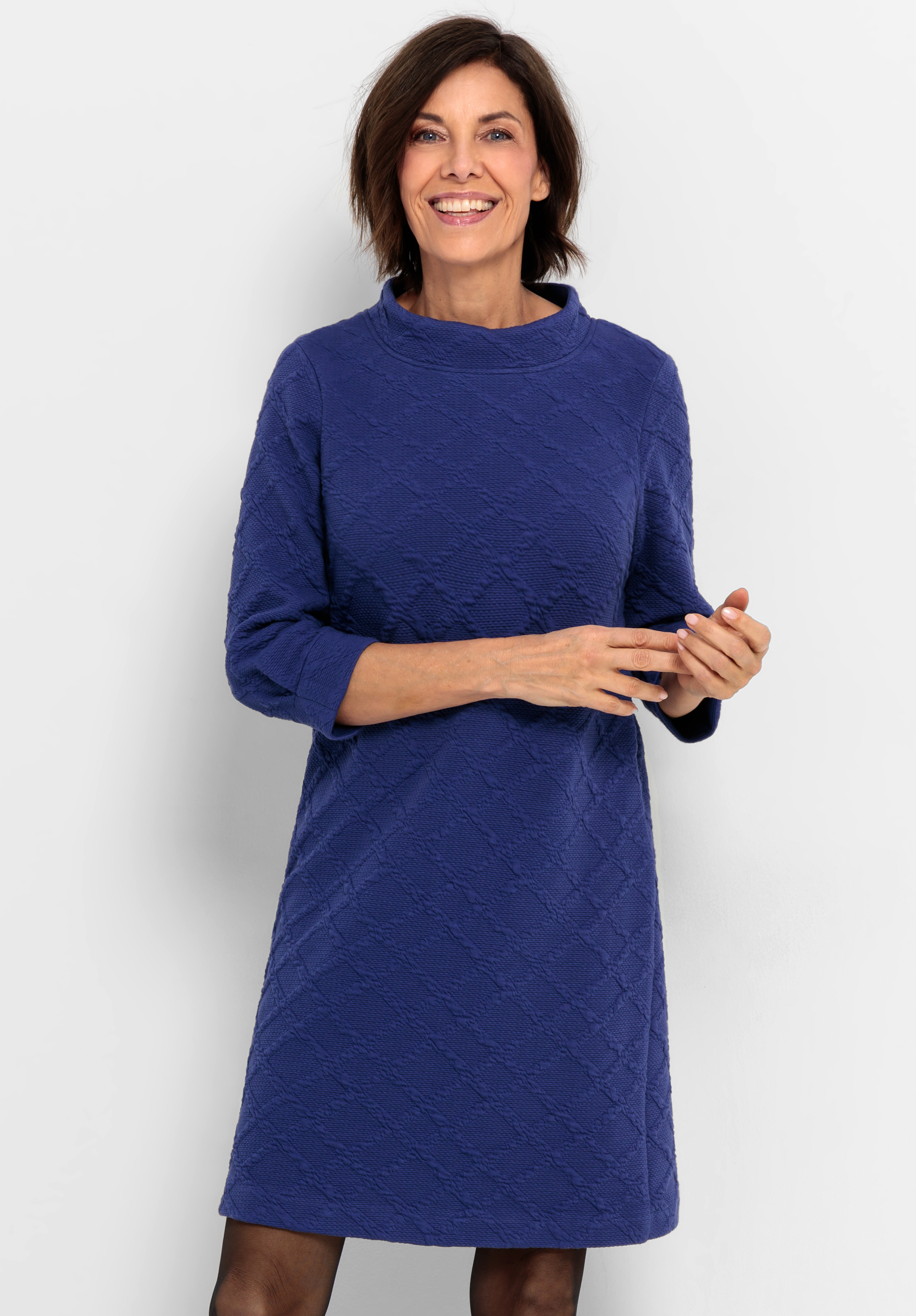 Jerseykleid ALICIA Eine Frau mit kurzen braunen Haaren, die ein strukturiertes, knielanges, langärmeliges blaues Kleid trägt, steht lächelnd vor einem schlichten weißen Hintergrund.