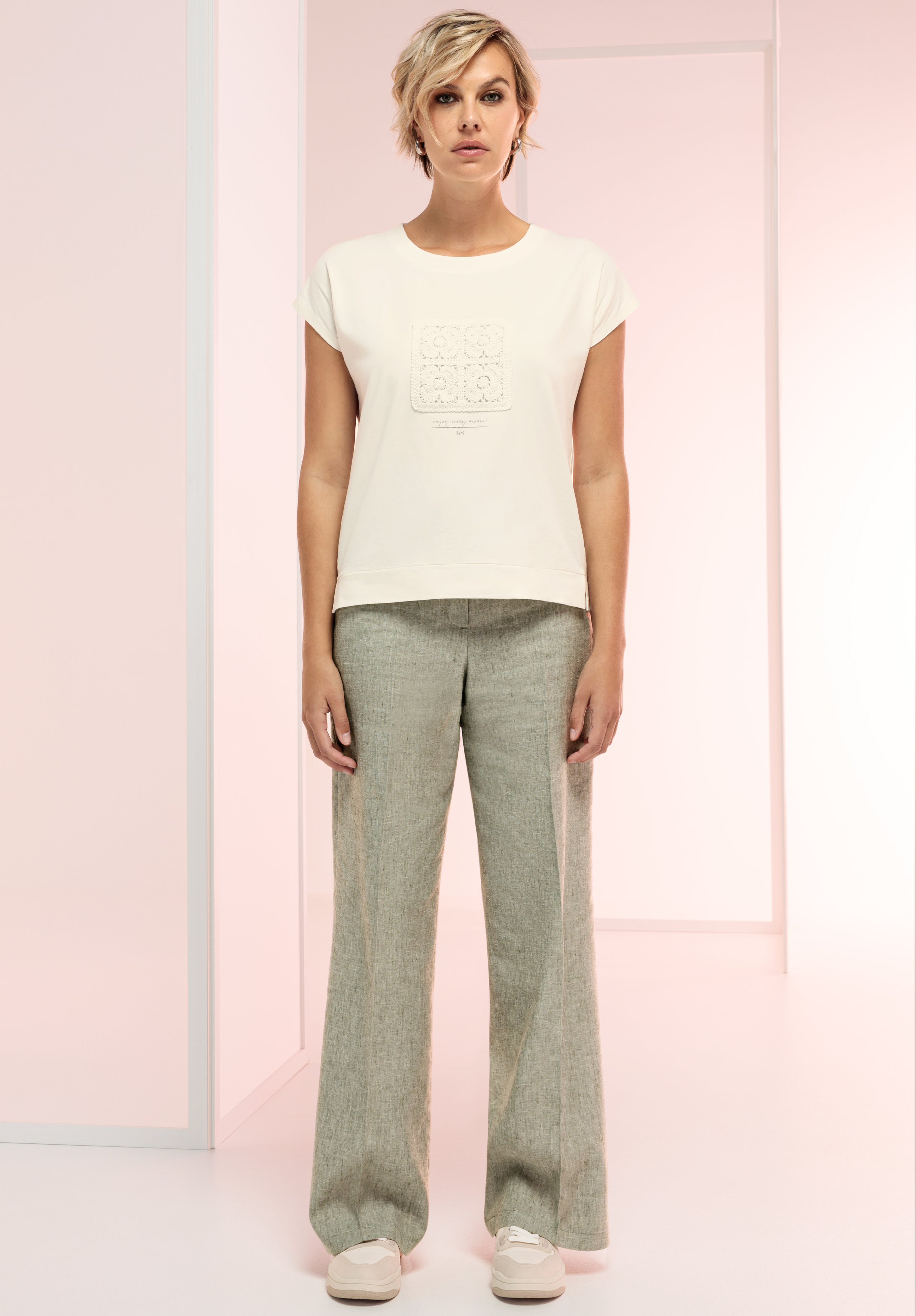 Eine Frau mit kurzen blonden Haaren steht nach vorne gerichtet. Sie trägt ein weißes T-Shirt mit dezentem Muster und eine locker sitzende, hellgraue Hose. Sie trägt weiße Turnschuhe in einem minimalistisch und sanft beleuchteten Raum.