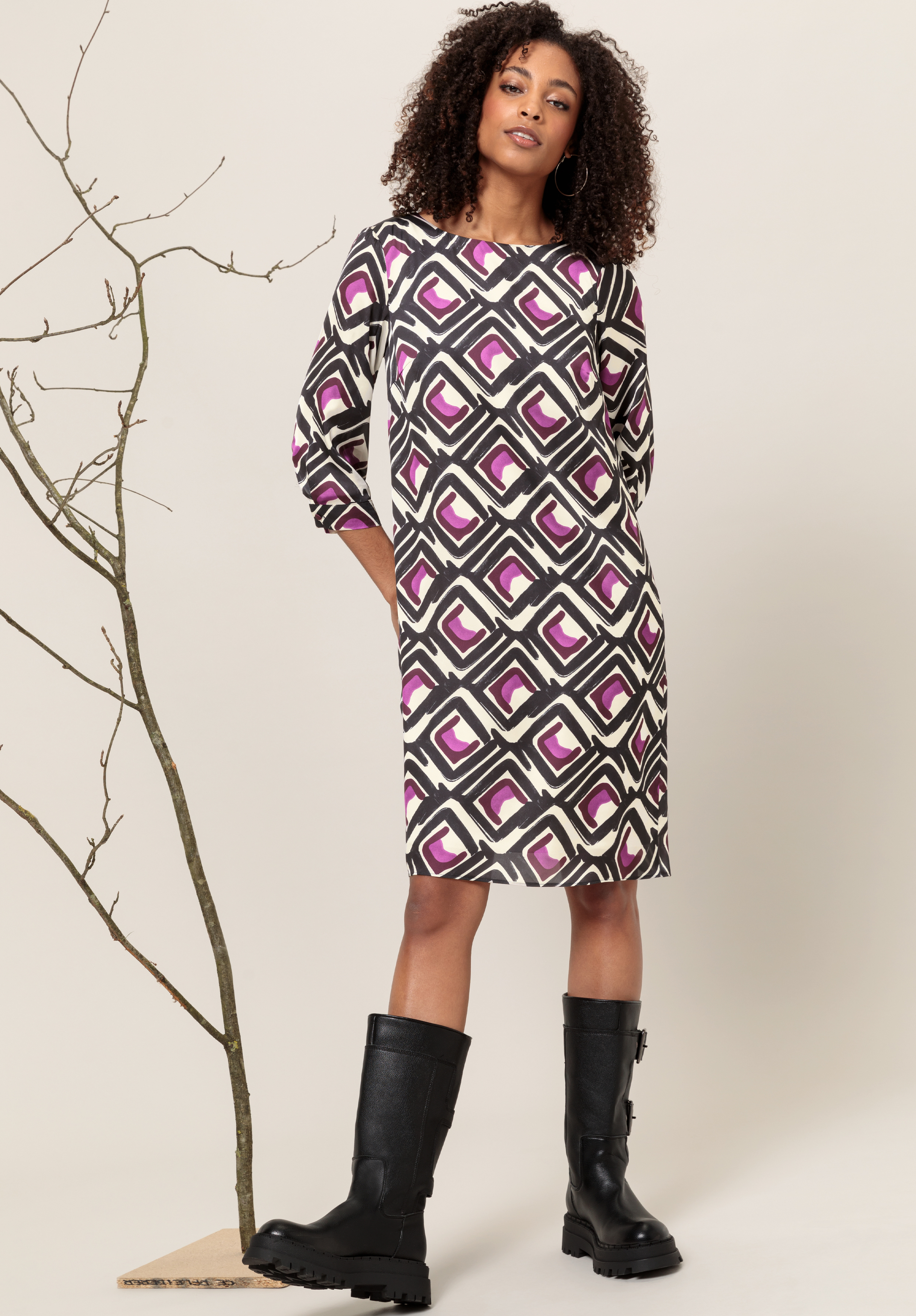 Eine Frau in einem knielangen Kleid mit einem auffälligen geometrischen Muster in Schwarz, Weiß und Lila steht neben einem kahlen Ast. Sie hat lockiges Haar und trägt schwarze kniehohe Stiefel.