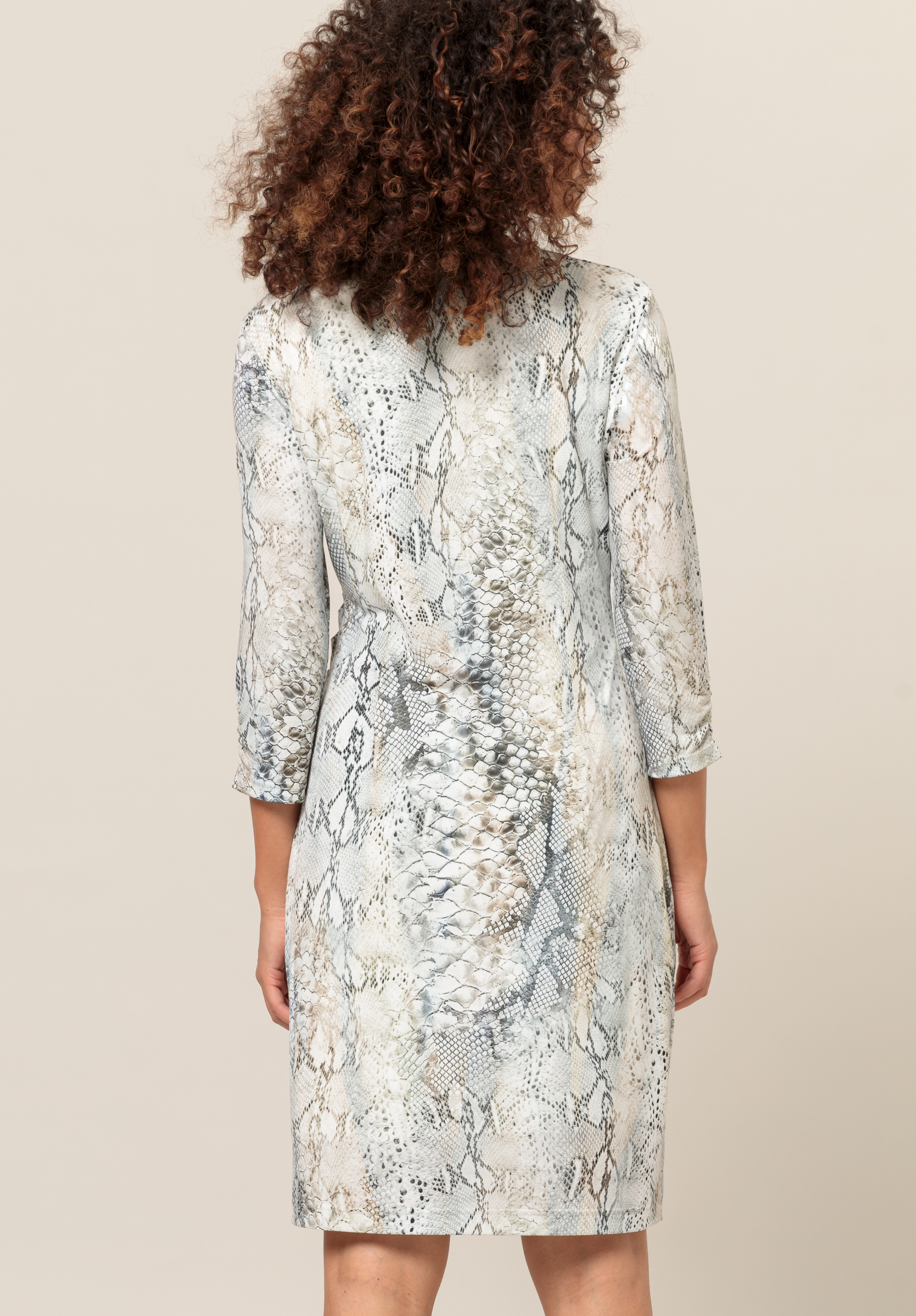 Von hinten ist eine Frau mit lockigem Haar zu sehen. Sie trägt ein knielanges, langärmeliges Kleid mit einem hellgrau-beigen Schlangenhautmuster. Der Hintergrund ist schlicht und neutral.