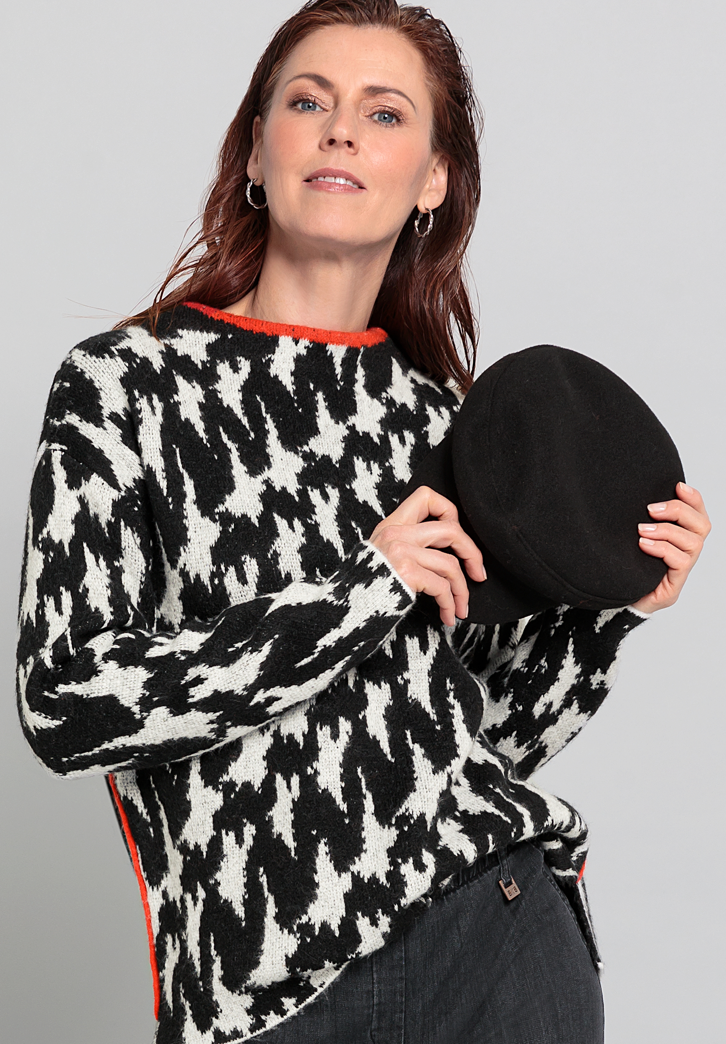 Eine Frau mit langen braunen Haaren, die einen schwarz-weiß gemusterten Pullover trägt, hält einen schwarzen Hut in der linken Hand. Vor einem schlichten grauen Hintergrund blickt sie selbstbewusst in die Kamera.