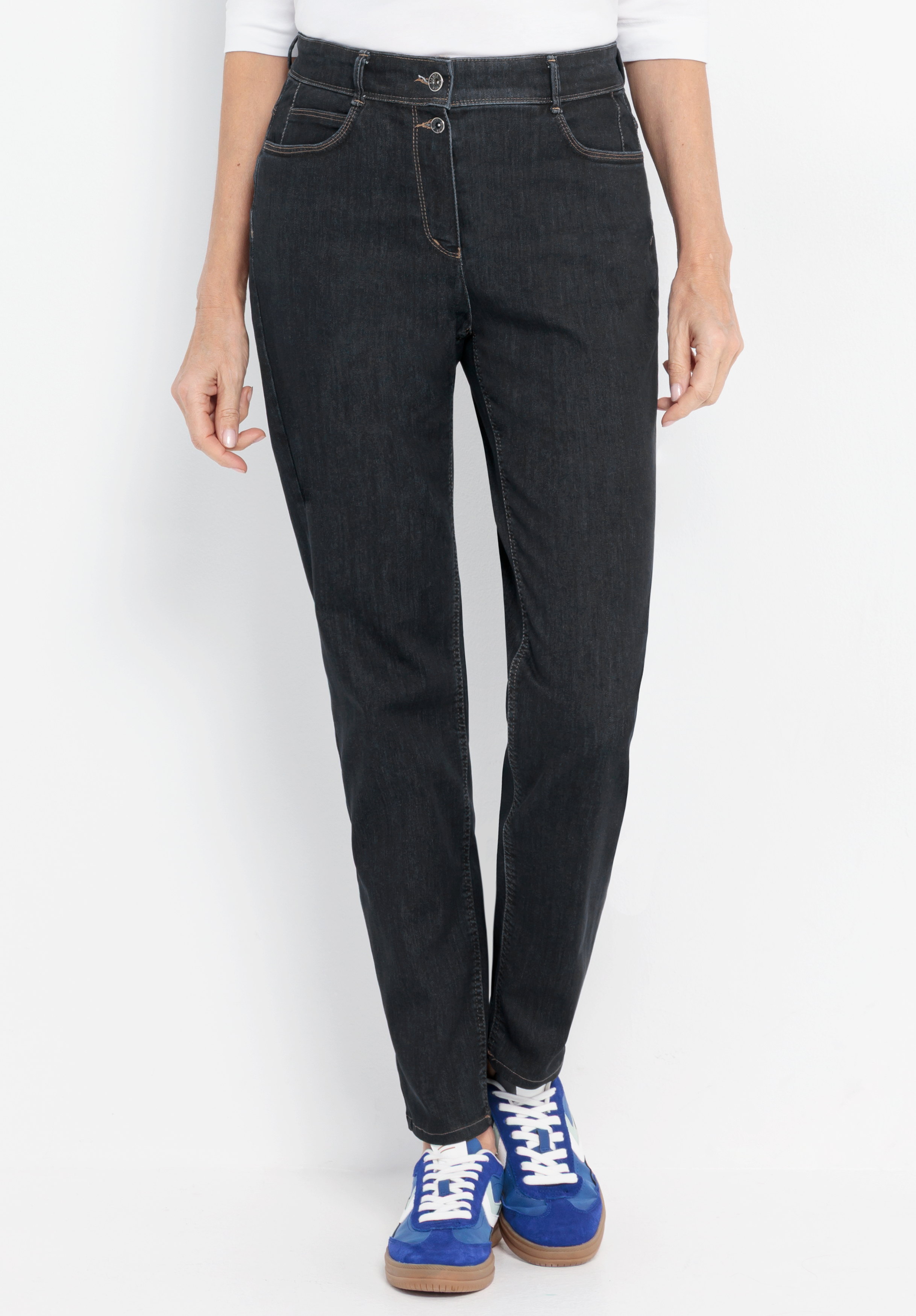 Eine Person, die eine dunkle, eng anliegende Jeans und blaue Turnschuhe trägt, steht vor einem weißen Hintergrund, die Hände locker an den Seiten. Zu sehen ist nur die untere Körperhälfte.