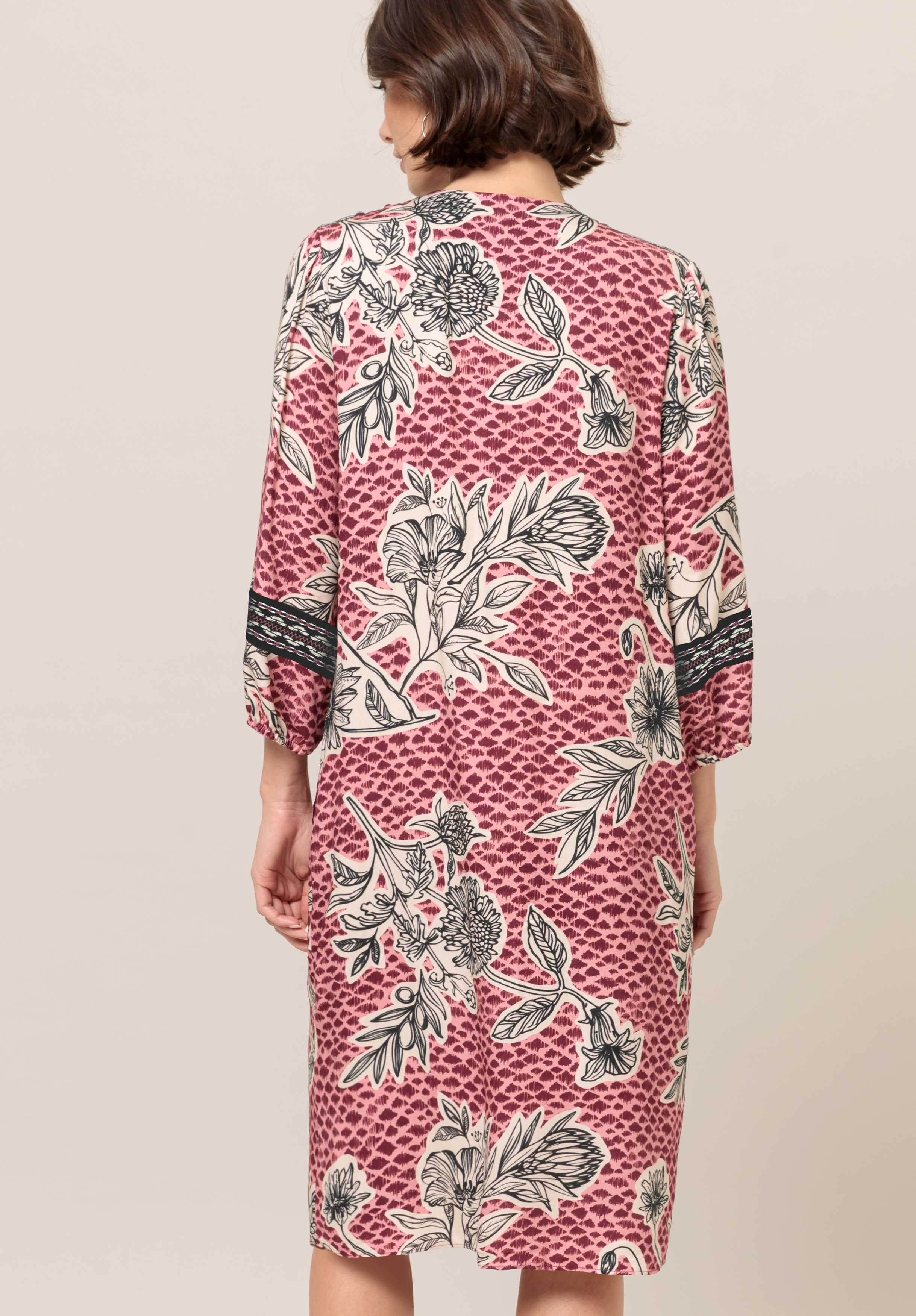 Von hinten ist eine Frau mit kurzen braunen Haaren zu sehen, die ein knielanges, langärmeliges Kleid mit rosa-weißem Blumenmuster und schwarzen Akzenten trägt.