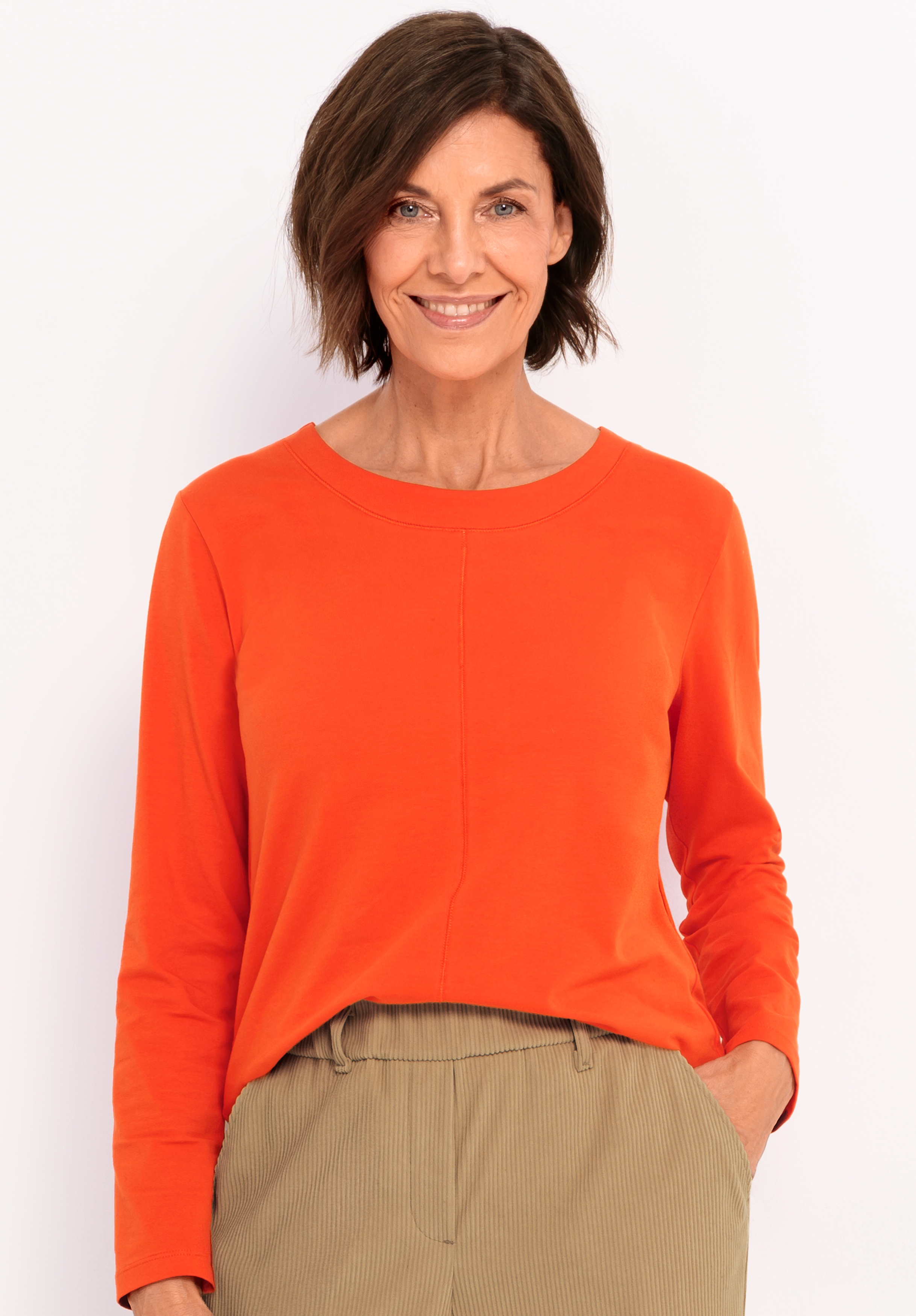 Rundhalsshirt DAMI Eine Frau mit kurzen braunen Haaren, die ein orangefarbenes langärmeliges Oberteil und eine beigefarbene Hose trägt, lächelt, während sie mit einer Hand in der Tasche vor einem schlichten weißen Hintergrund steht.