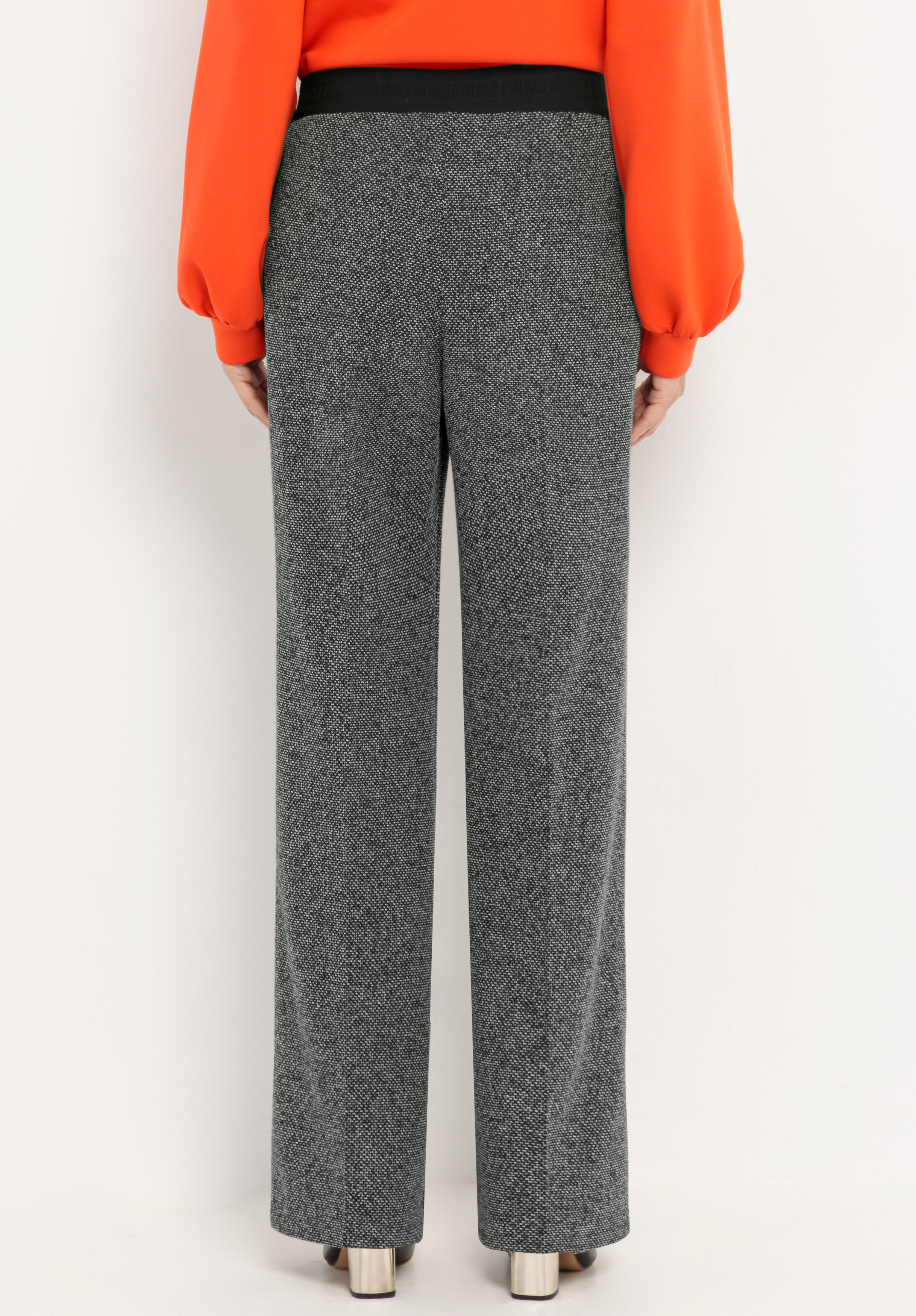 Vor einem schlichten weißen Hintergrund steht eine Person mit dem Rücken zur Kamera. Sie trägt eine graue, strukturierte Hose, ein leuchtend orangefarbenes Langarm-Oberteil und Schuhe mit Metallic-Absatz.