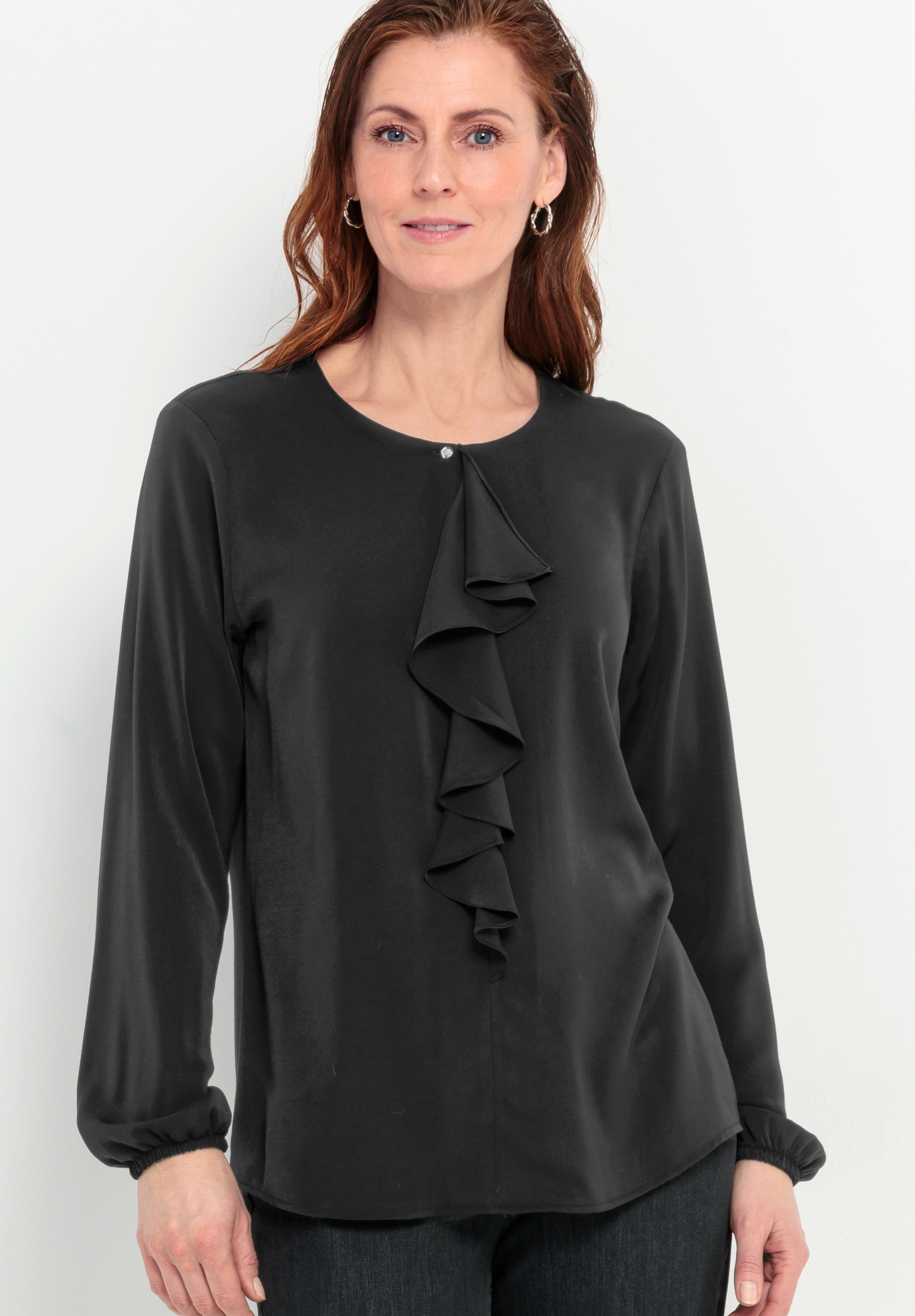 Eine Frau mit langen, braunen Haaren trägt eine schwarze, langärmelige Bluse mit Rüschenverzierung an der Vorderseite und einem kleinen Knopf am Ausschnitt. Sie steht vor einem schlichten weißen Hintergrund und lächelt sanft.