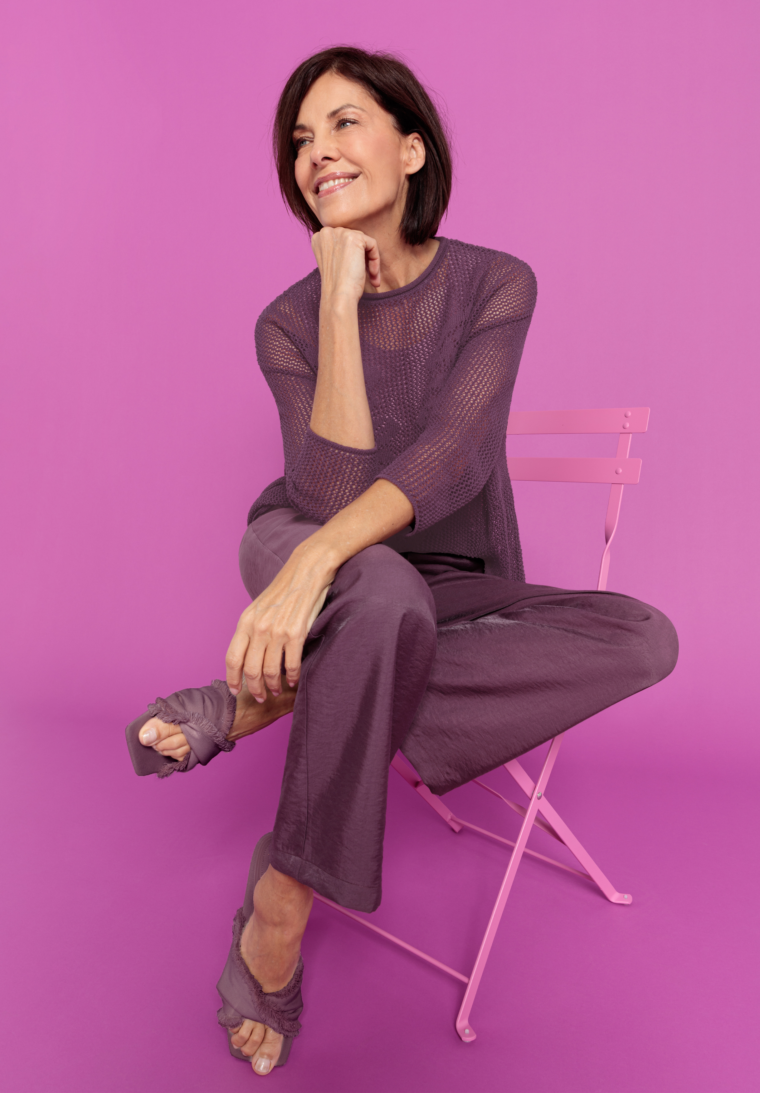 Eine lächelnde Frau mit kurzen braunen Haaren sitzt auf einem hellrosa Stuhl vor einem rosafarbenen Hintergrund und trägt ein lilafarbenes Netzoberteil, eine passende Hose und offene Schuhe mit Stoffriemen.