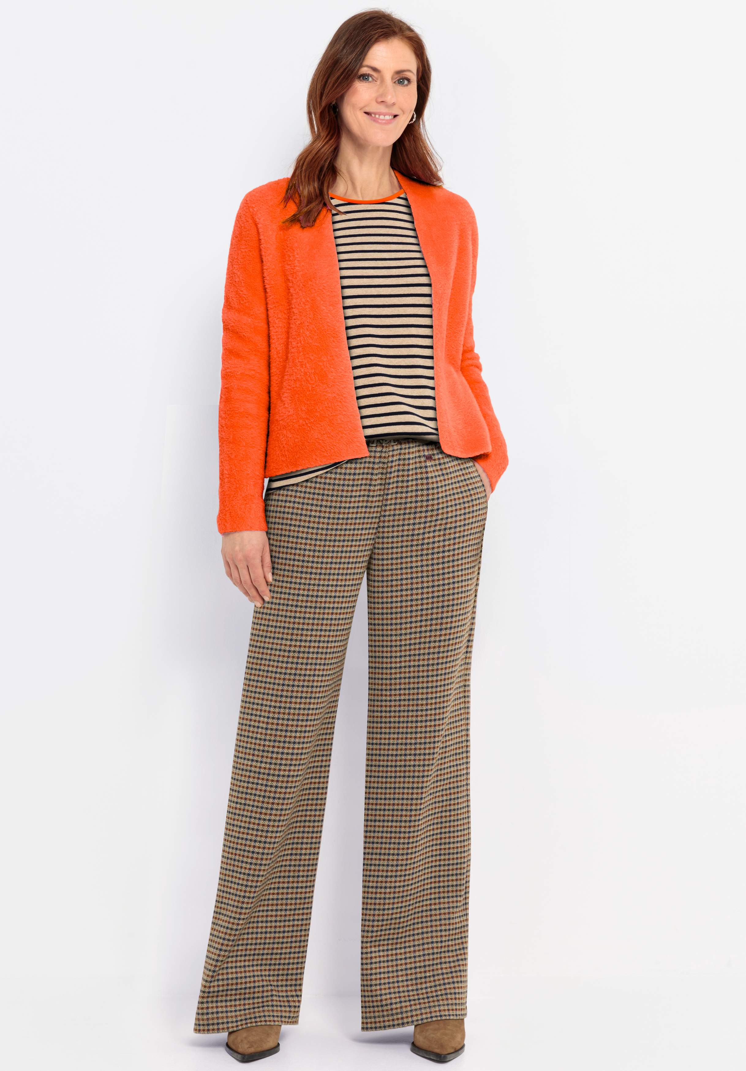 Eine Frau steht vor einem weißen Hintergrund und trägt eine orangefarbene strukturierte Jacke, ein schwarz-beige gestreiftes Oberteil und eine braun karierte Hose mit weitem Bein und braunen Absätzen. Sie lächelt und hat gewelltes braunes Haar.