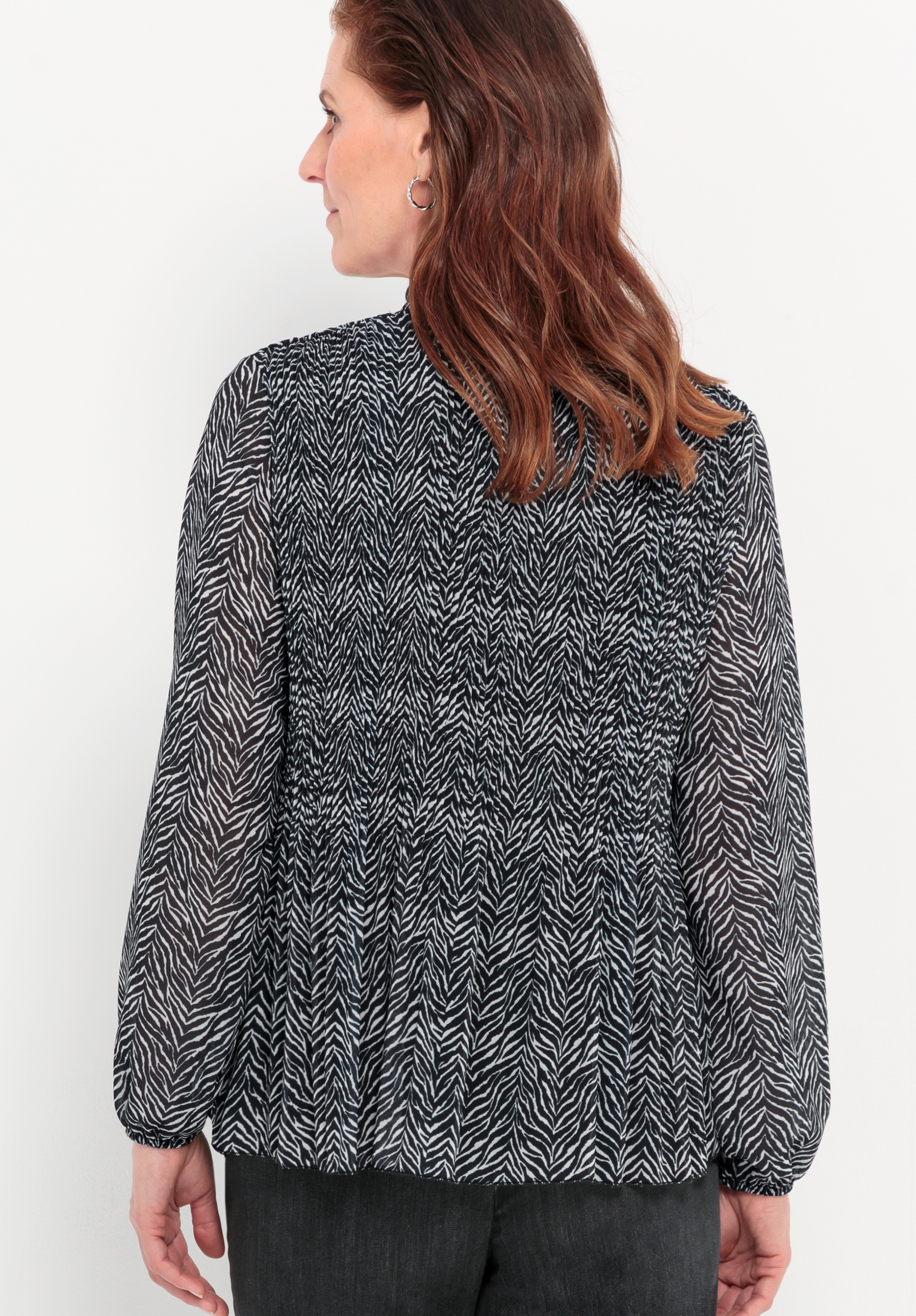 Eine Frau mit welligem braunem Haar ist von hinten zu sehen; sie trägt eine langärmelige, schwarz-weiß gemusterte Bluse mit dezentem Zebradruck und eine schwarze Hose. Der Hintergrund ist einfarbig und hell.
