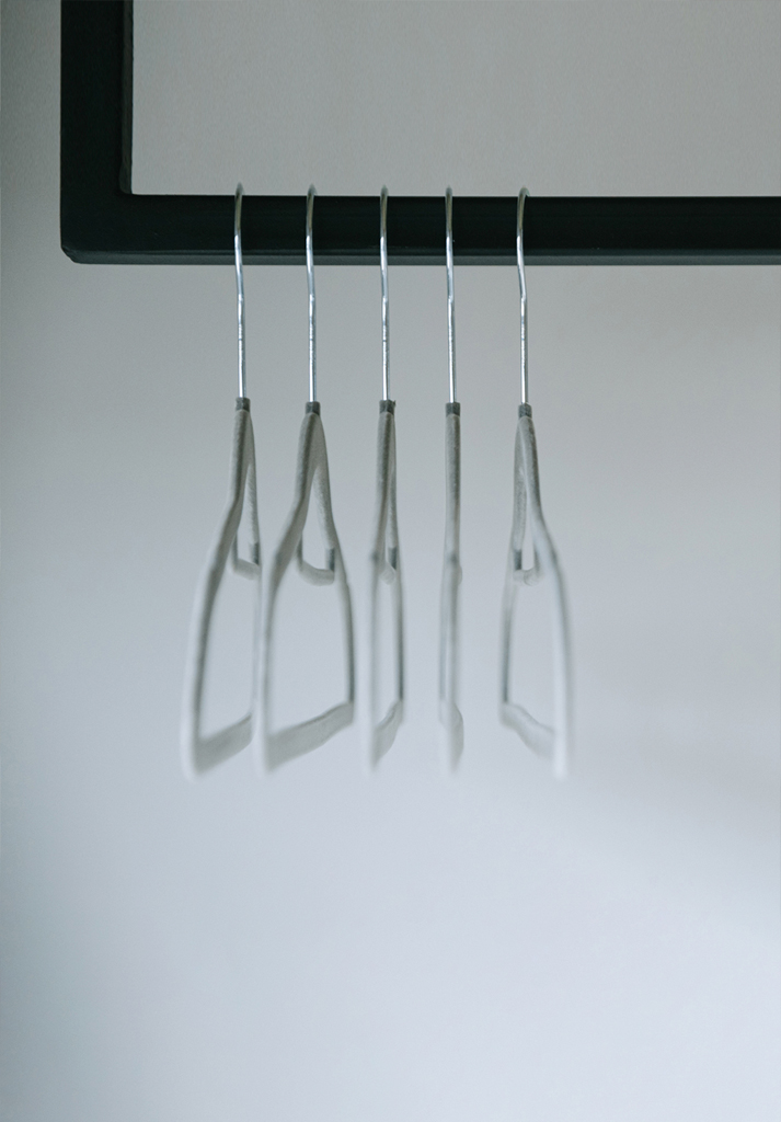 Fünf leere Kleiderbügel hängen an einem schwarzen Kleiderständer vor einem schlichten, hellen Hintergrund. Das Bild wirkt minimalistisch und leicht verschwommen.