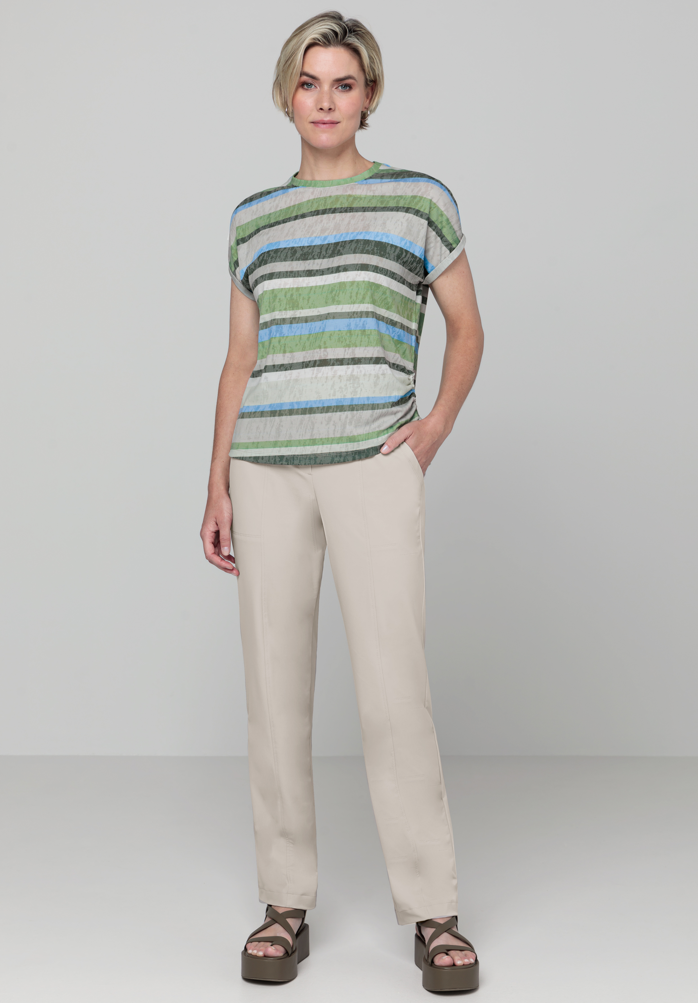 Vor einem schlichten Hintergrund steht eine Person, die ein kurzärmeliges, grün-blau gestreiftes Oberteil, eine beige Hose und braune Plateausandalen trägt, eine Hand in der Tasche hat und einen neutralen Gesichtsausdruck hat.