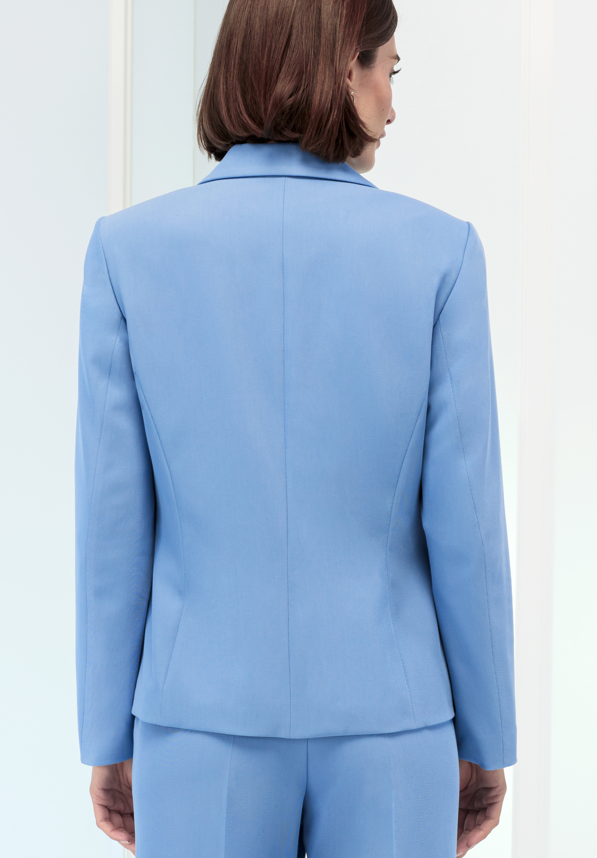 Eine Frau mit glattem braunem Haar ist von hinten zu sehen. Sie trägt ein hellblaues Jackett und eine dazu passende Hose und steht in einem Innenbereich vor einem hellen Hintergrund.