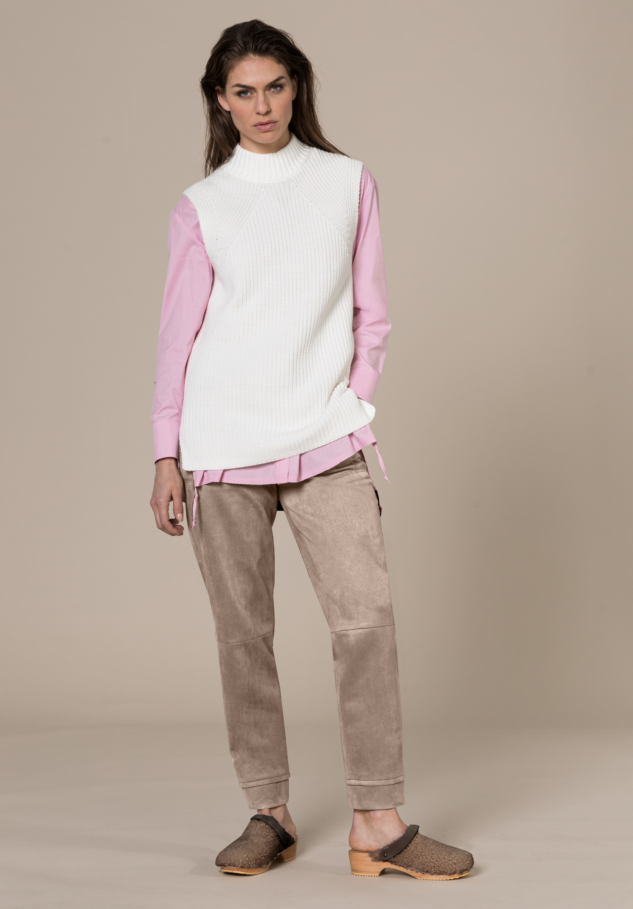 Eine Frau steht vor einem beigen Hintergrund. Sie trägt ein langes rosa Hemd unter einer ärmellosen weißen Strickweste, taupefarbene Hosen und braune Slipper. Sie hat lange Haare und einen neutralen Gesichtsausdruck.
