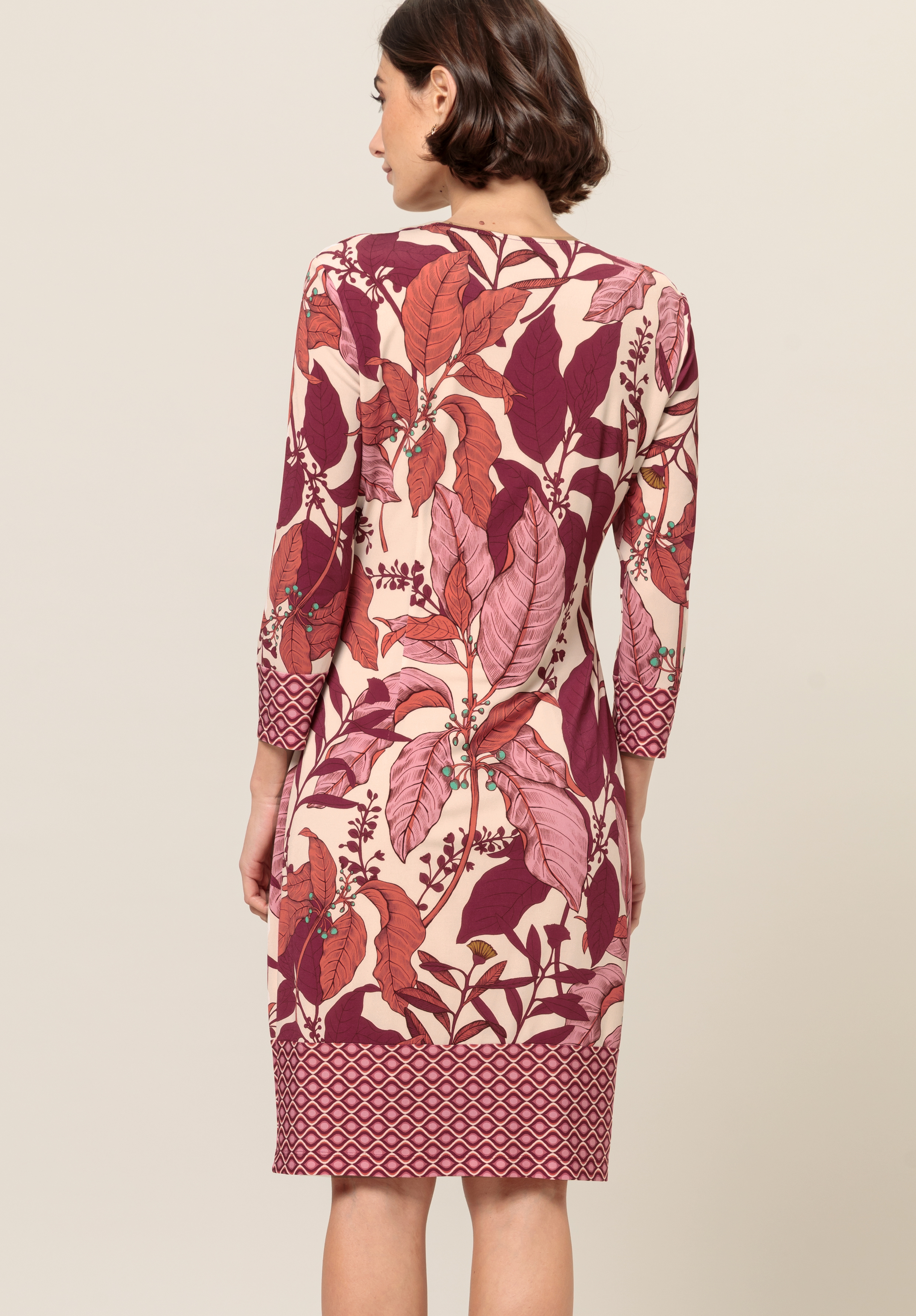 Eine Frau mit kurzen braunen Haaren steht mit abgewandtem Gesicht da und trägt ein knielanges Kleid mit Dreiviertelärmeln. Das Kleid weist ein kräftiges, blattartiges Blumenmuster in Rosa-, Burgunder- und Beigetönen auf.