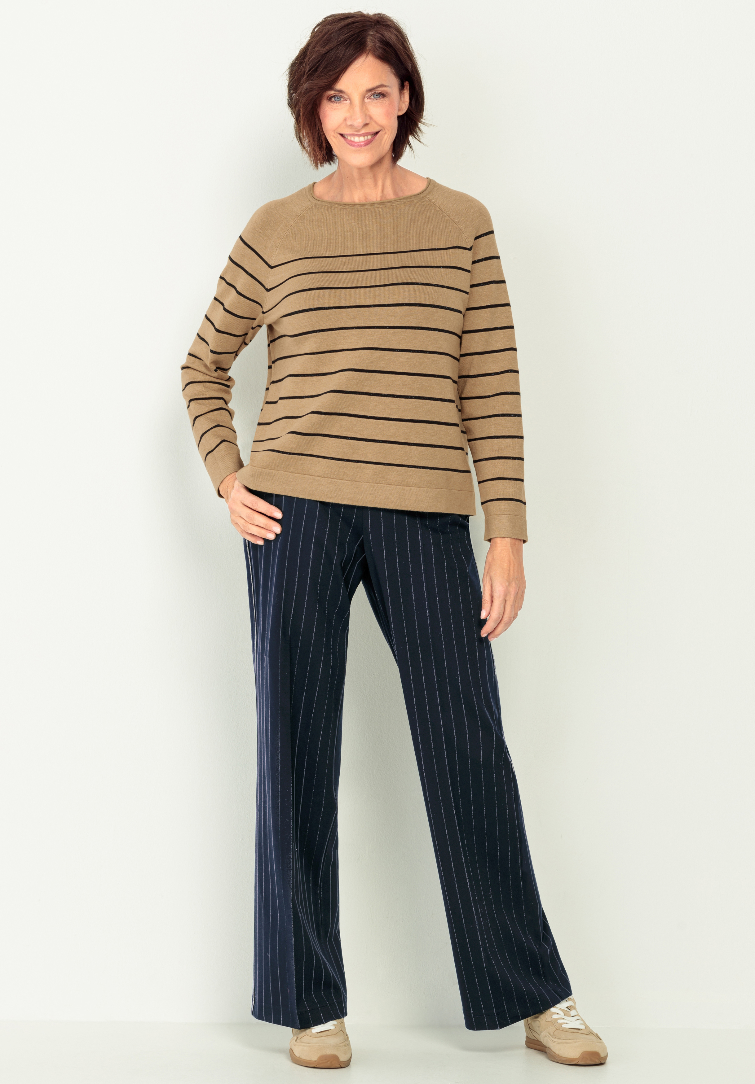 Eine Frau lächelt, während sie einen hellbraunen Pullover mit schwarzen Querstreifen, eine marineblaue Hose mit Nadelstreifen und beigefarbene Turnschuhe trägt. Sie steht vor einem schlichten, hellen Hintergrund und hat eine Hand in ihrer Tasche.